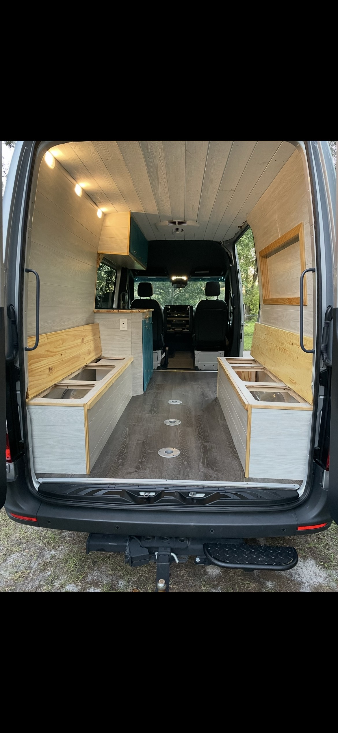 Rear view of a modern RV rental camper van interior. Features light wood paneling, benches with storage, and a small kitchen area.. Mercedes-Benz Sprinter 2019