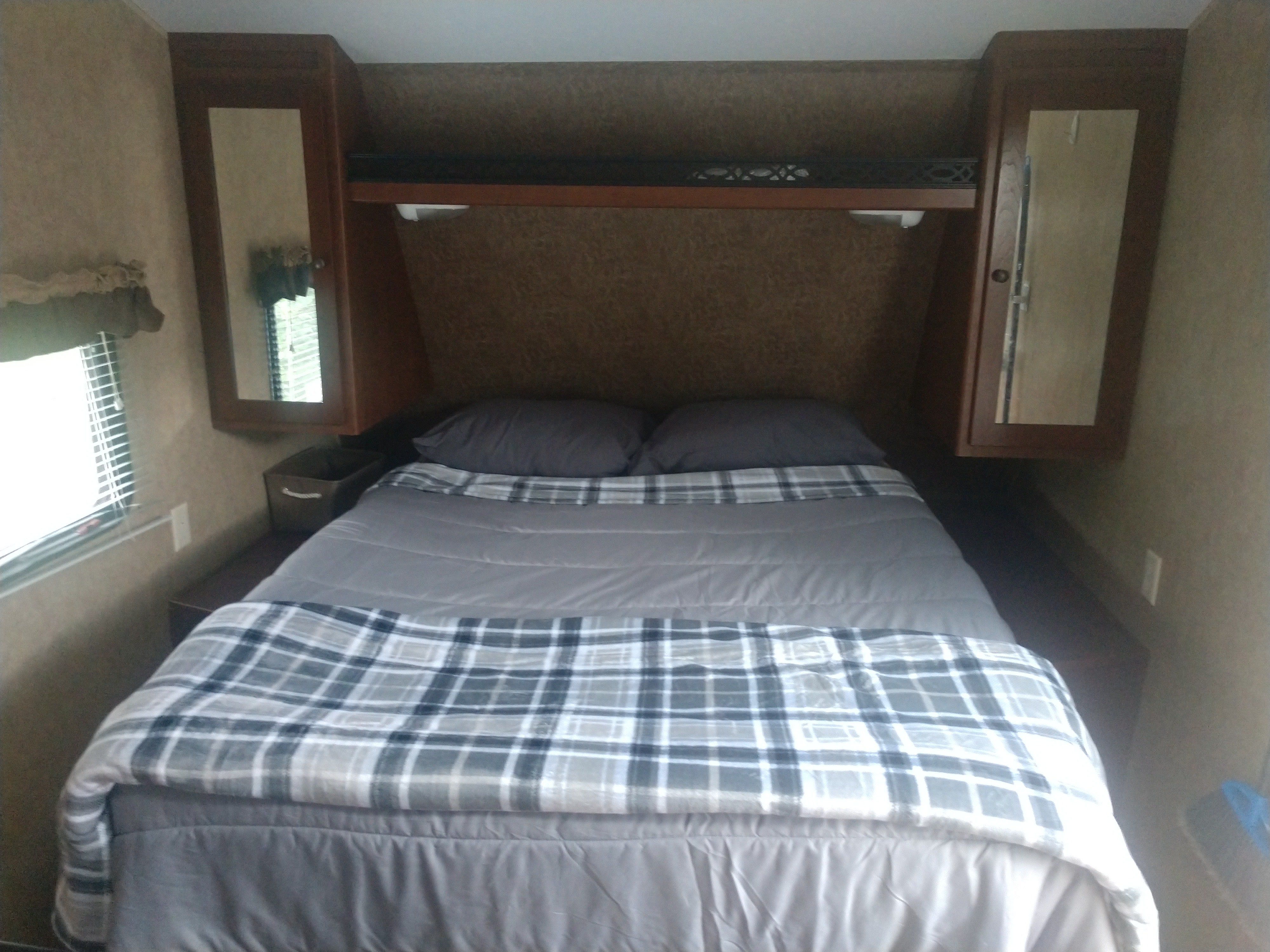 Cozy RV rental bedroom featuring a made bed with grey bedding, plaid blanket, side window, and mirrored overhead storage.. Aspen Trail Aspen Trail Trailer 2013