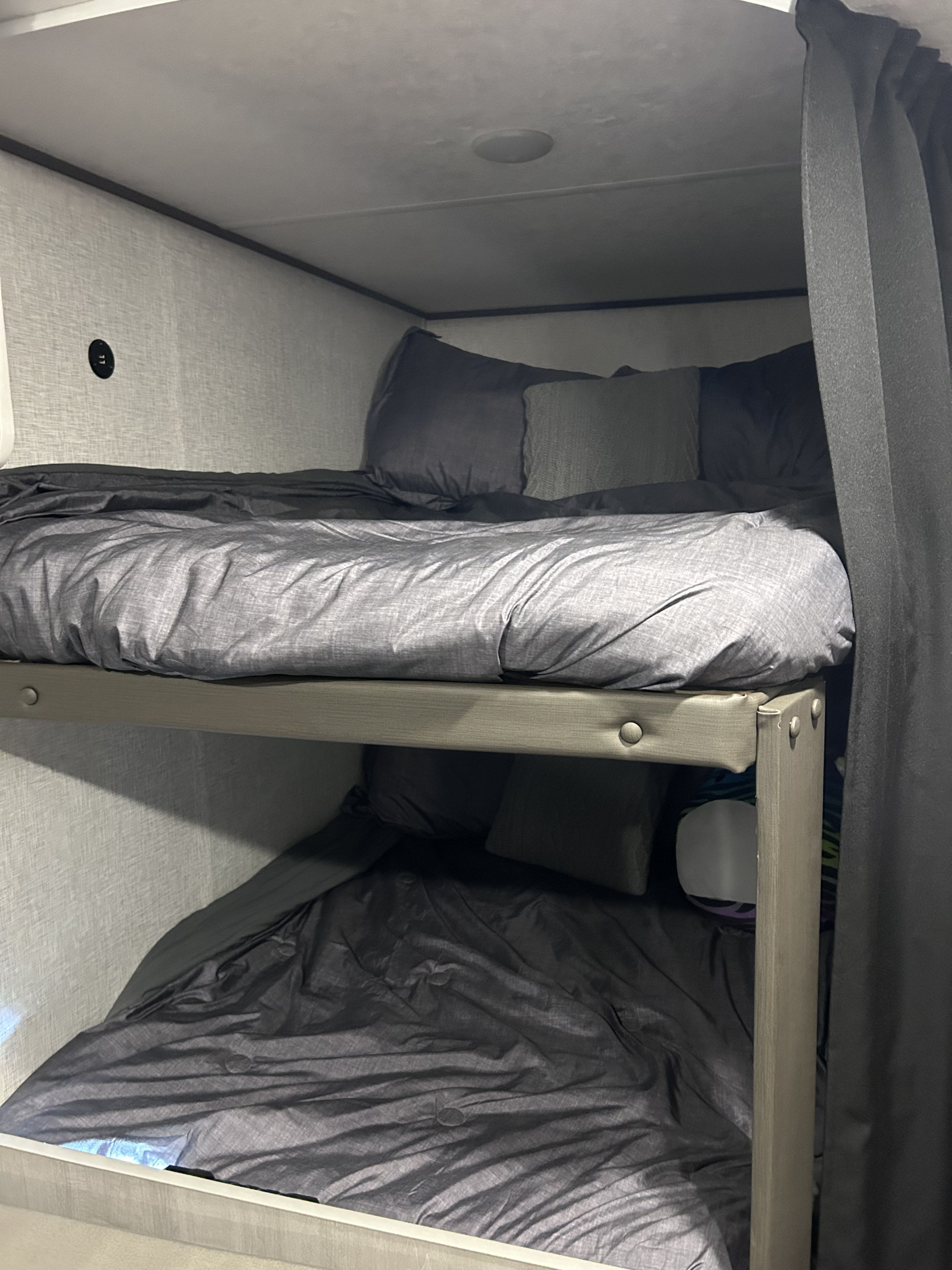 Cozy grey bunk beds in an RV rental with bedding, pillows, and a wall outlet for charging. Dark curtains offer privacy.. Crossroads Zinger Lite 2025