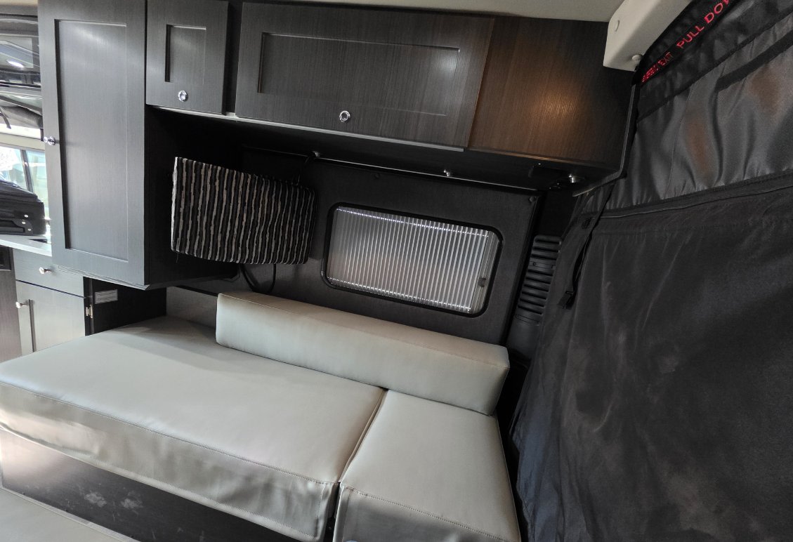 Inside an rv rental: dark cabinets, comfy light-colored bench/bed, small window with blinds, and emergency exit curtain.. Roadtrek Chase Class B 2022