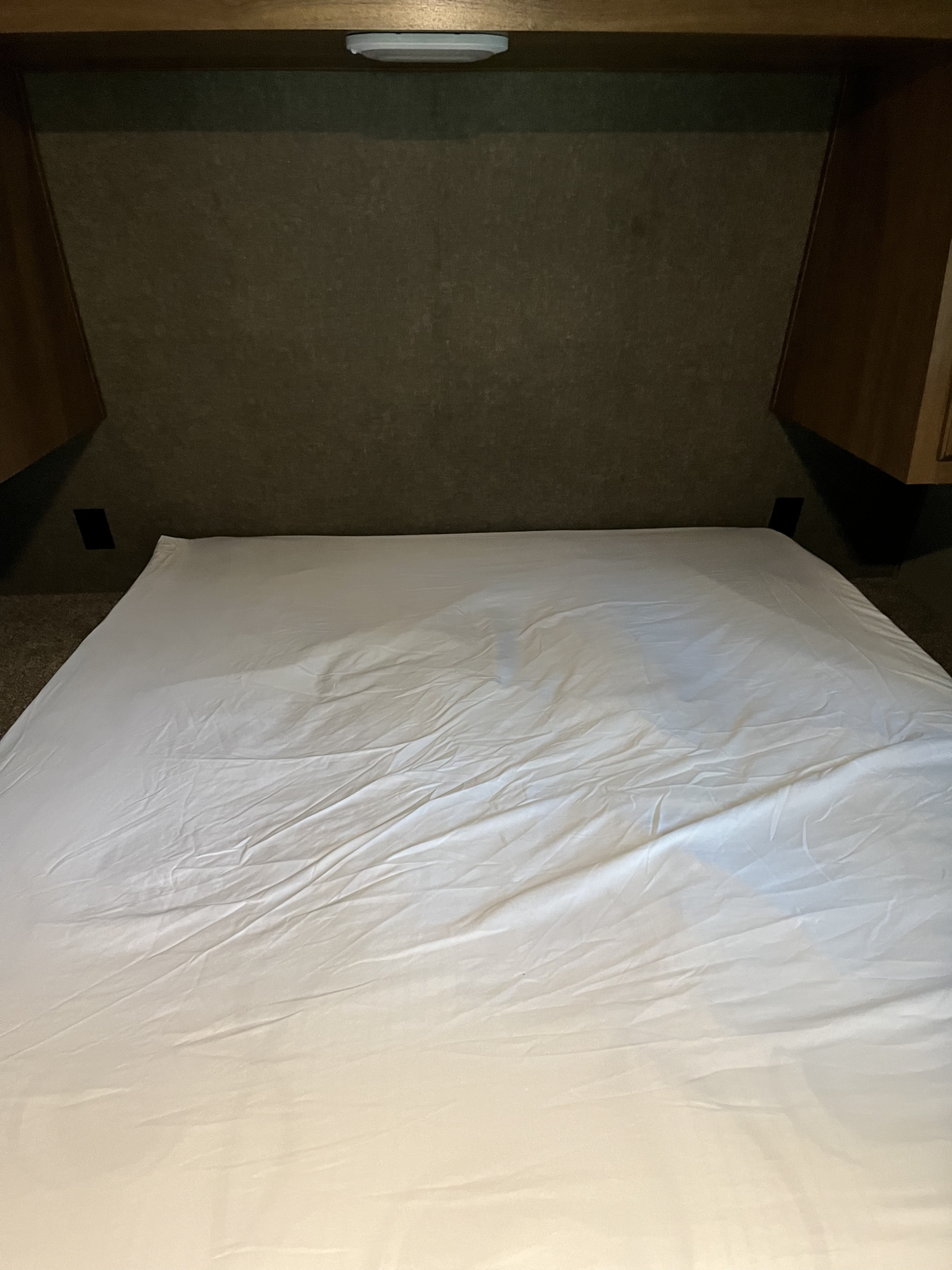 Inside an RV rental, a compact bedroom with a bed, white sheets, and built-in amenities like outlets and an overhead light.. Keystone RV Springdale 2019