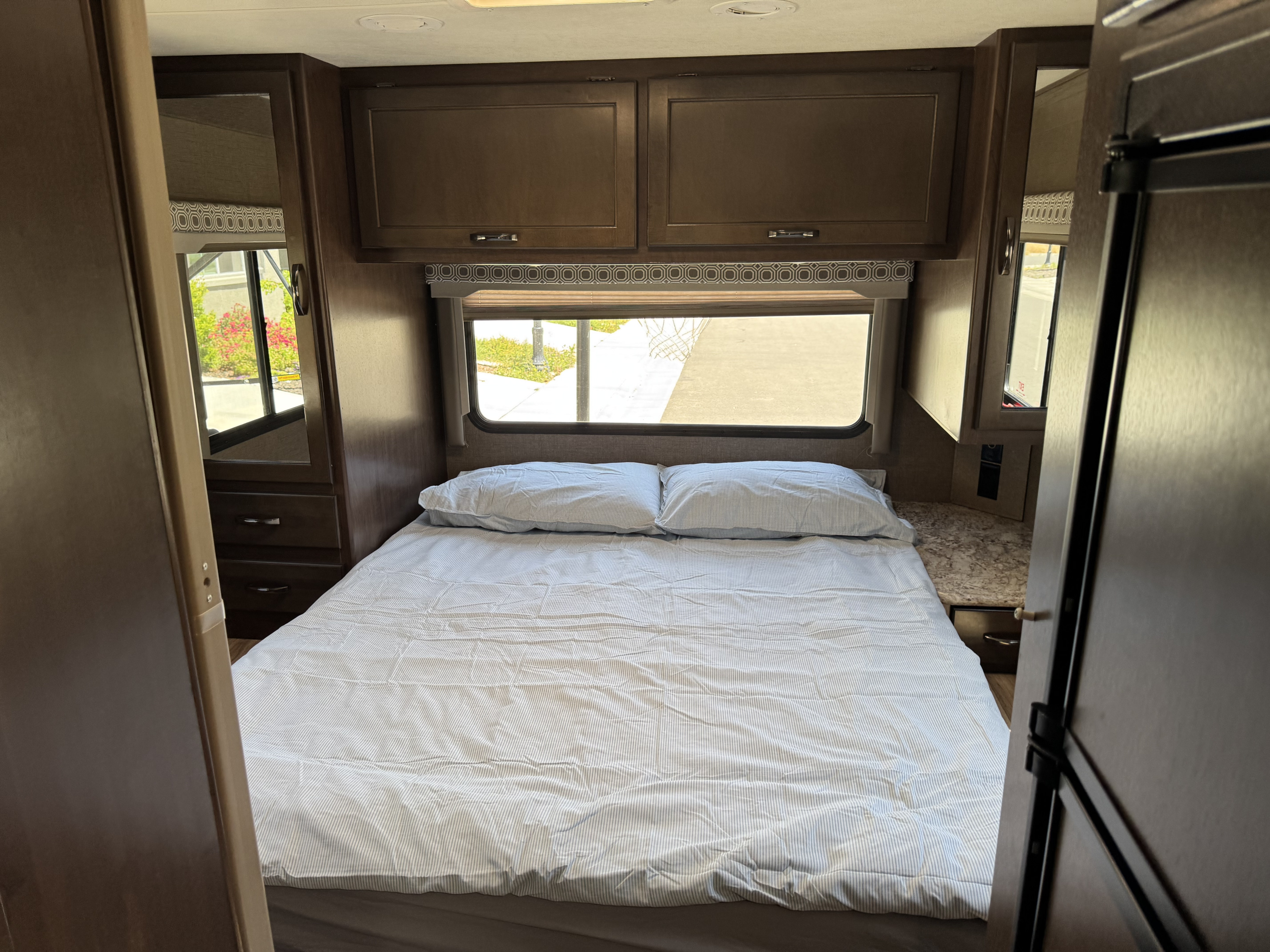 This RV rental bedroom offers a cozy queen bed with striped linens, storage cabinets, and a window looking outdoors.. Thor Four Winds 2020