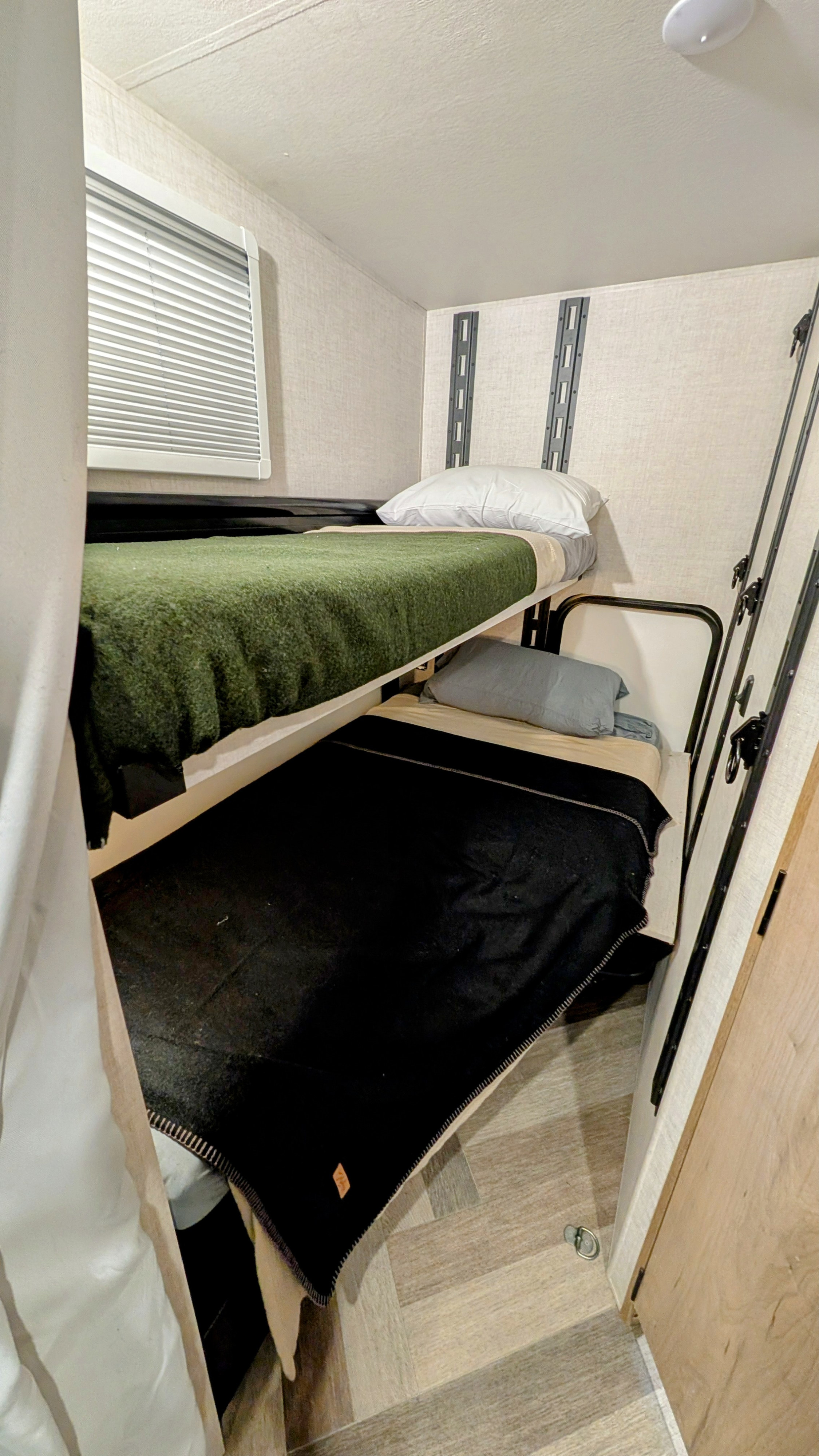 Cozy bunk beds inside an RV rental feature green and black blankets, a window, and wall tracks on wood-look flooring.. Ember Overland 2023