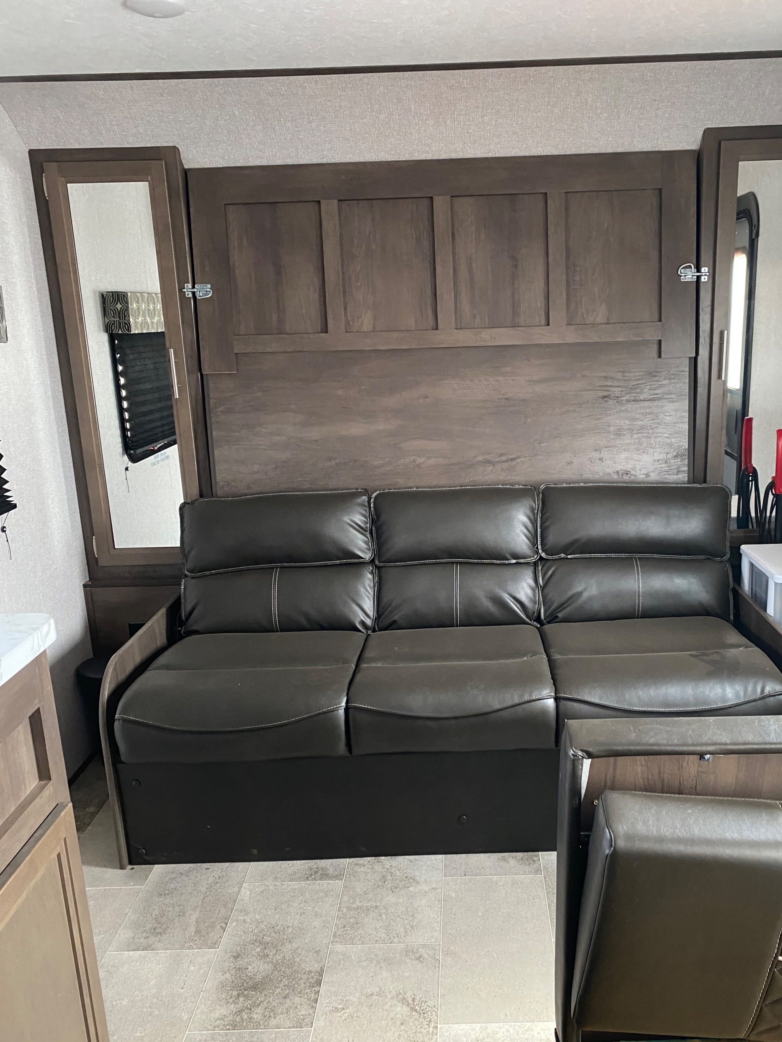 Inside an RV rental, a dark brown sofa with light stitching sits against a wood-paneled wall, possibly a murphy bed.. Dutchmen Coleman LX 1705RB 2021