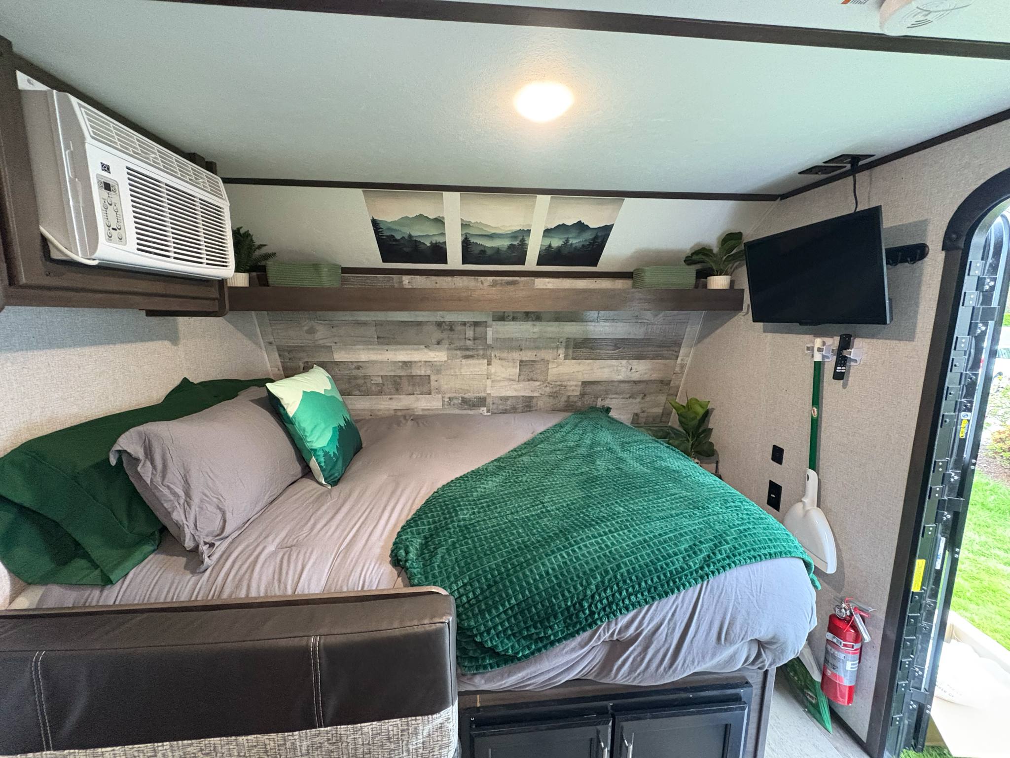 Cozy RV rental bedroom features a comfortable bed with green accents, wall-mounted AC, TV, and scenic mountain art.. Coleman Lantern 17B 2021