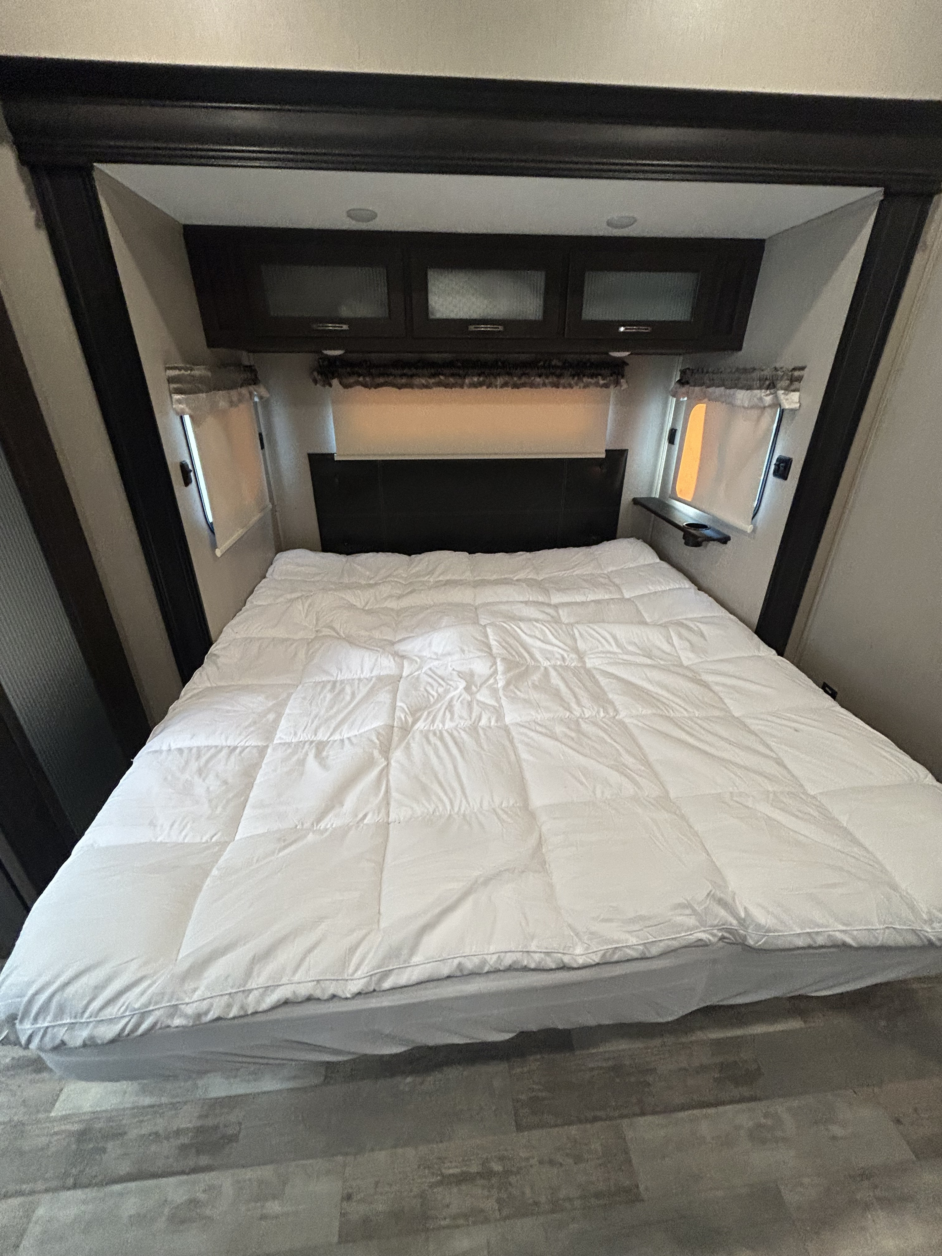 A cozy RV rental bedroom features a white-covered bed, overhead cabinets, and small windows for comfortable travel.. Forest River Coachmen Brookstone 2021