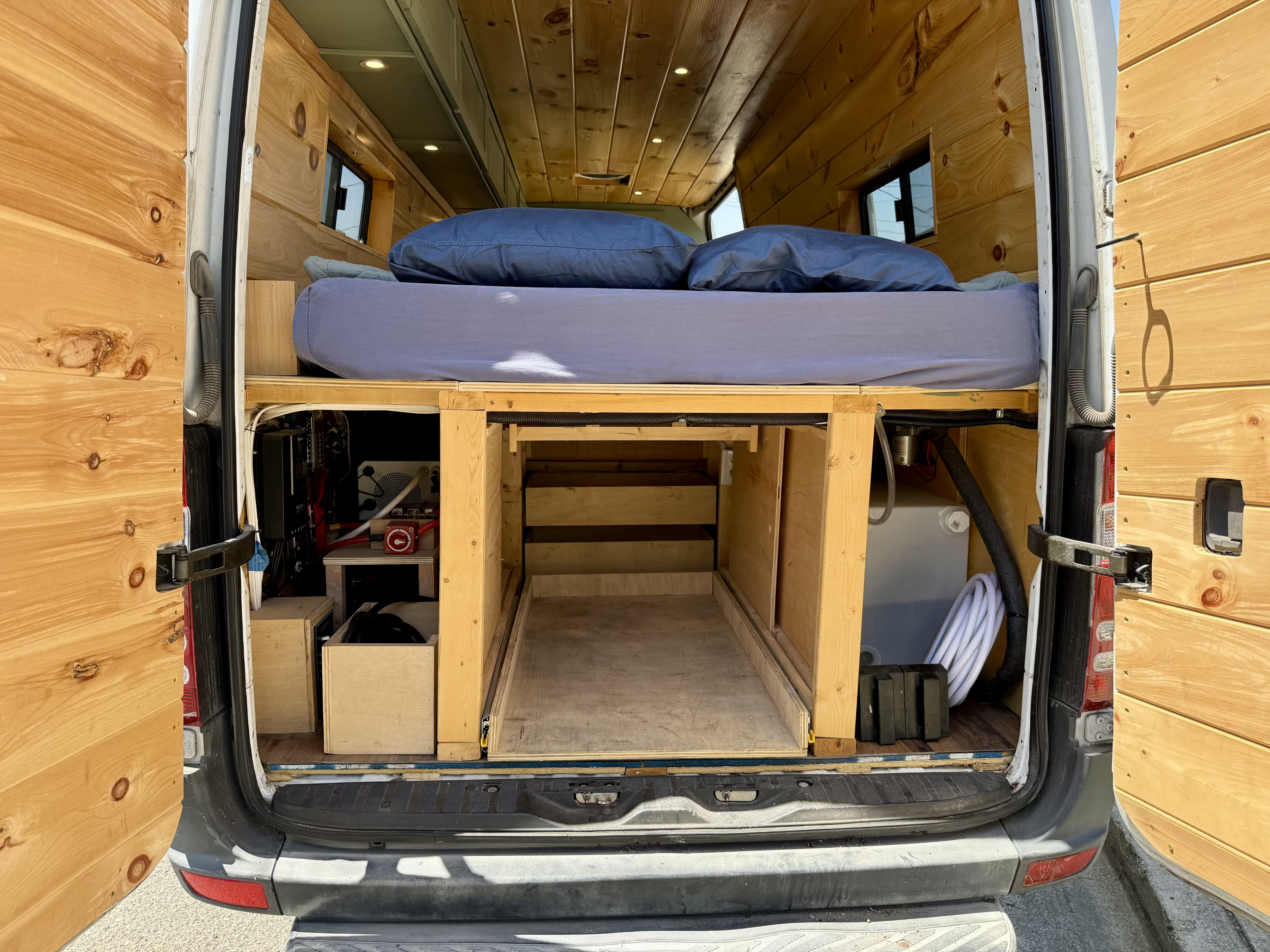 View of a custom rv rental van's rear interior, showing a wooden bed platform, pull-out storage, and utilities.. Dodge Sprinter 3500 2007