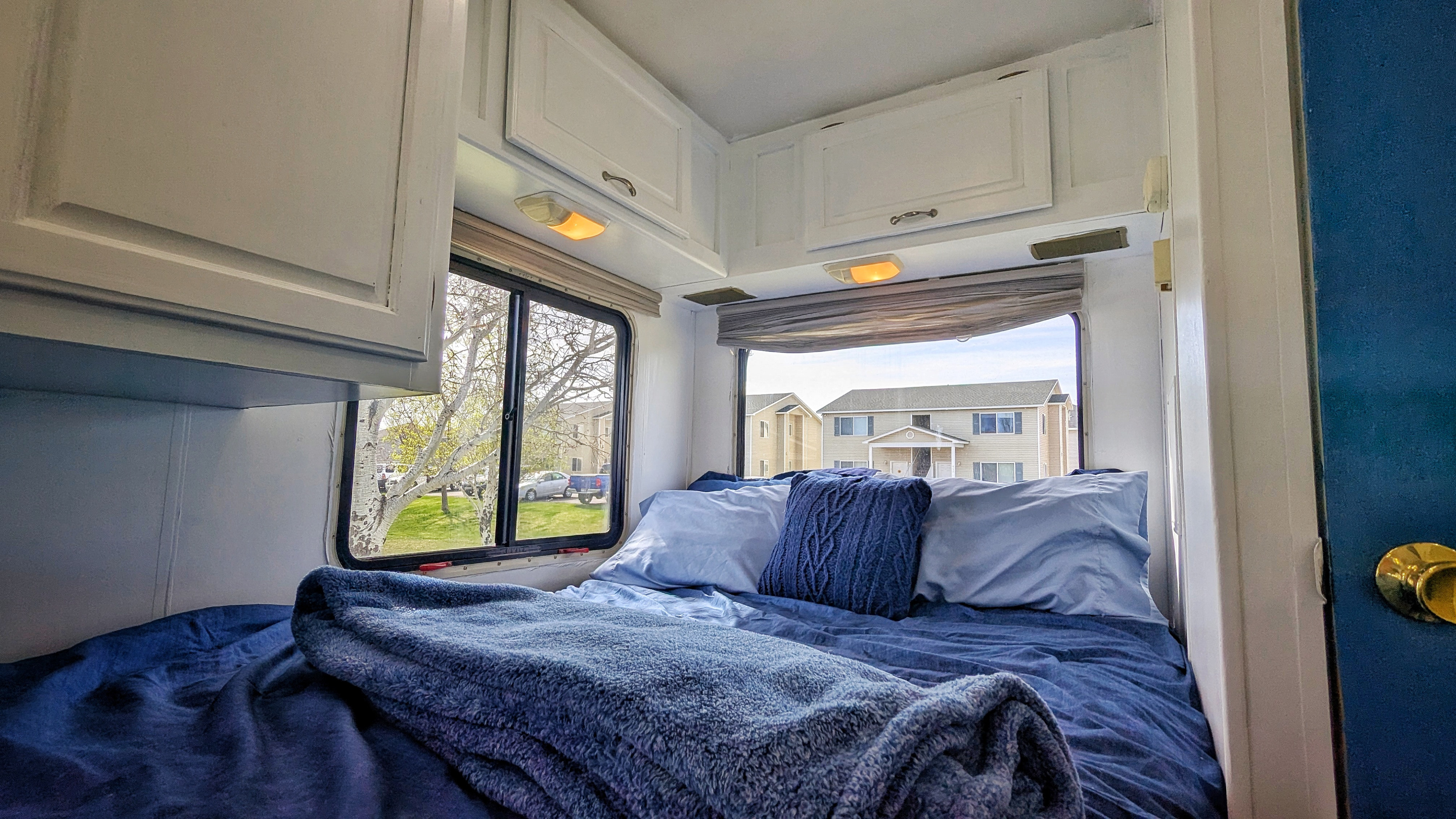 Cozy RV rental bedroom featuring a bed with blue bedding, overhead cabinets, and windows offering a view of the outside.. Four Winds Majestic 2004