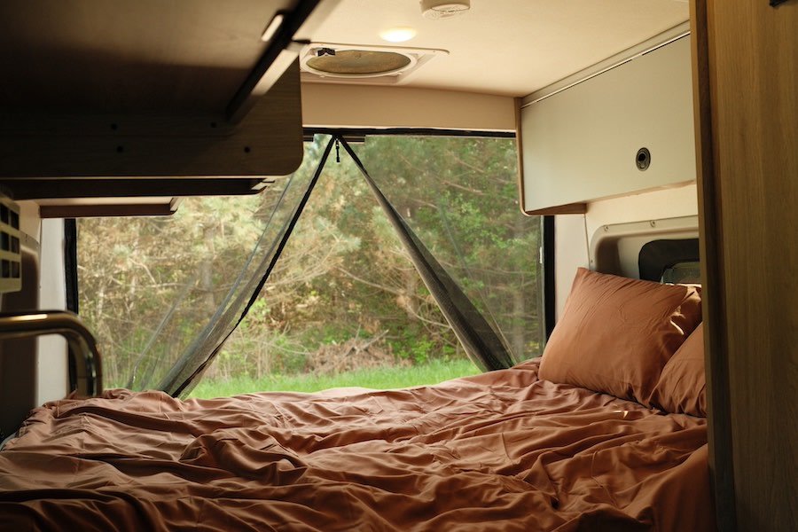 Inside an RV rental: a cozy bed with brown bedding, offering a view of a serene forest through a protective mesh screen.. Winnebago Solis 2022