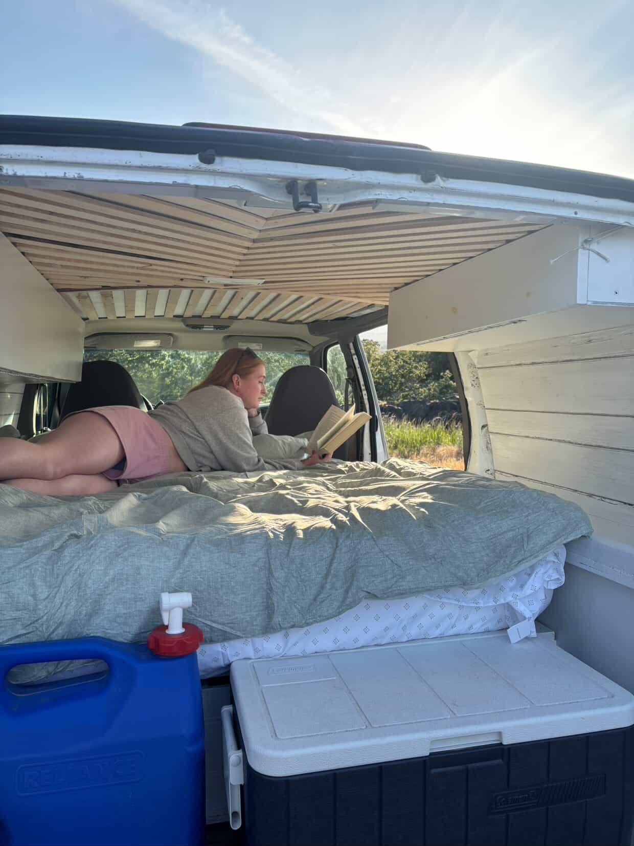 Person relaxing in an **rv rental** van, reading on a comfy bed. The custom interior features wooden ceiling slats and storage.. Gmc Custom 2003