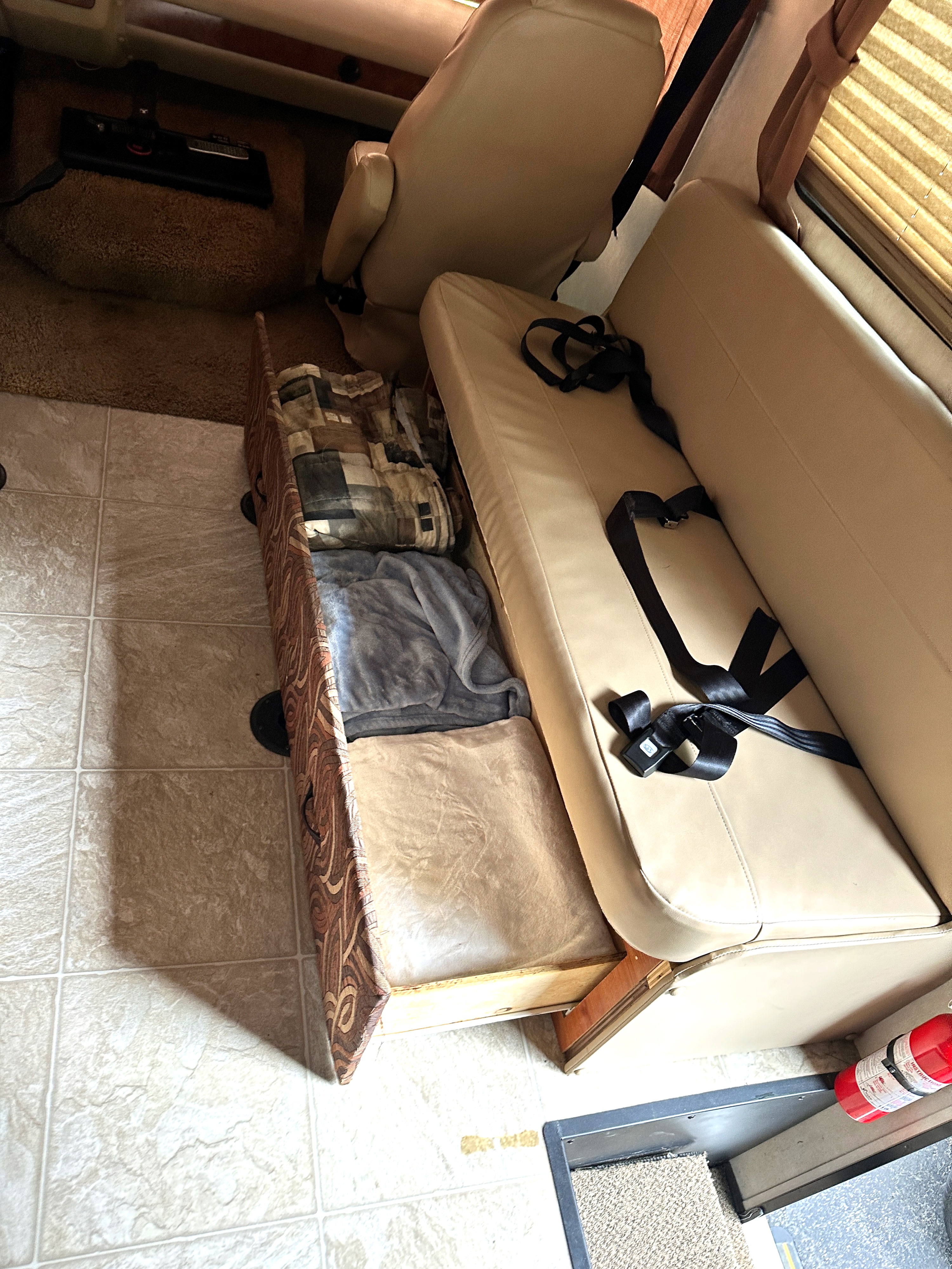 RV rental interior: Beige convertible couch with seatbelts, open storage with bedding, tiled floor, and front seats.. Coachmen Mirada 2009