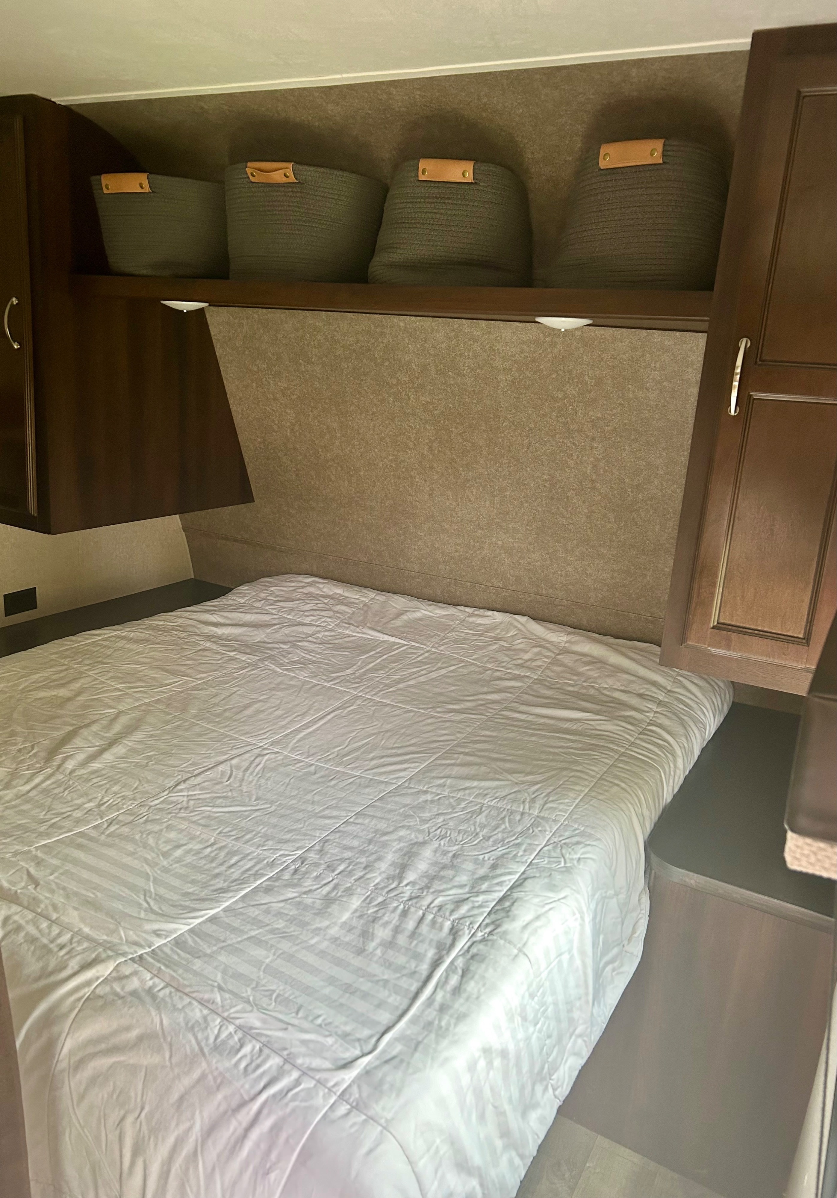Cozy RV rental bedroom features a white striped bed, dark wood overhead cabinets, and woven storage baskets.. Jayco Jay Flight SLX 2018