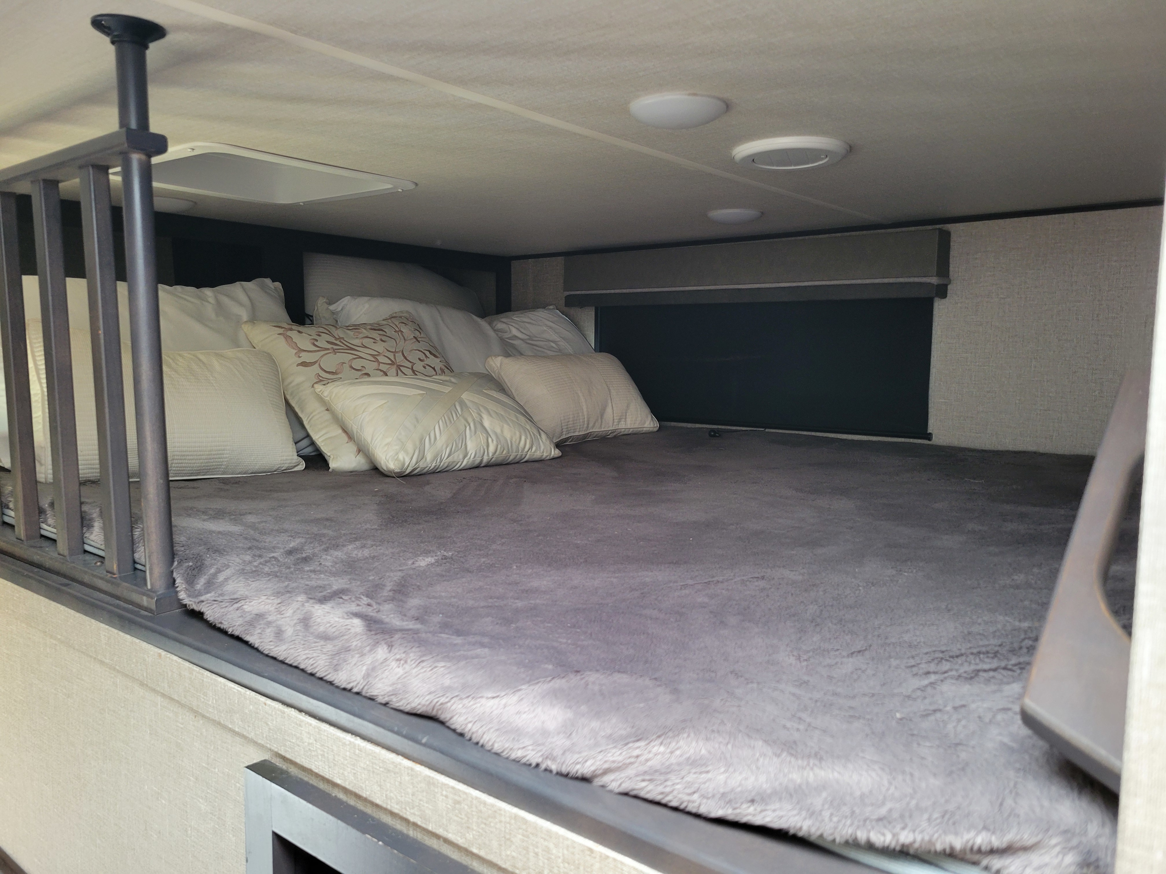 Cozy loft bed in an RV rental, with a fluffy grey blanket, pillows, ceiling lights, and a safety railing.. Grand Design Reflection 2020