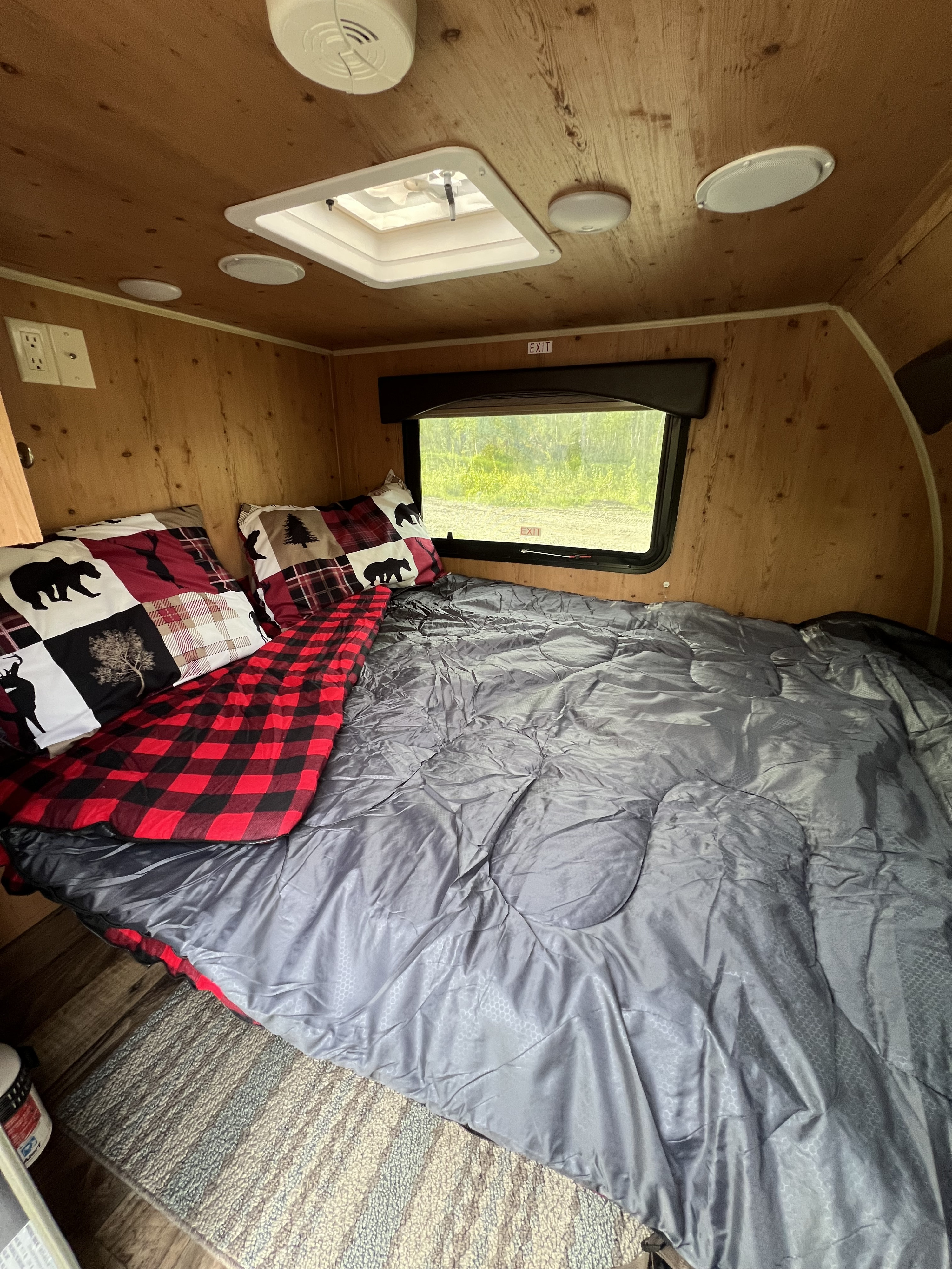 This RV rental offers a cozy interior with a bed, bear-themed pillows, red plaid blanket, and a nature view window.. Sunset Park RV 109 2018
