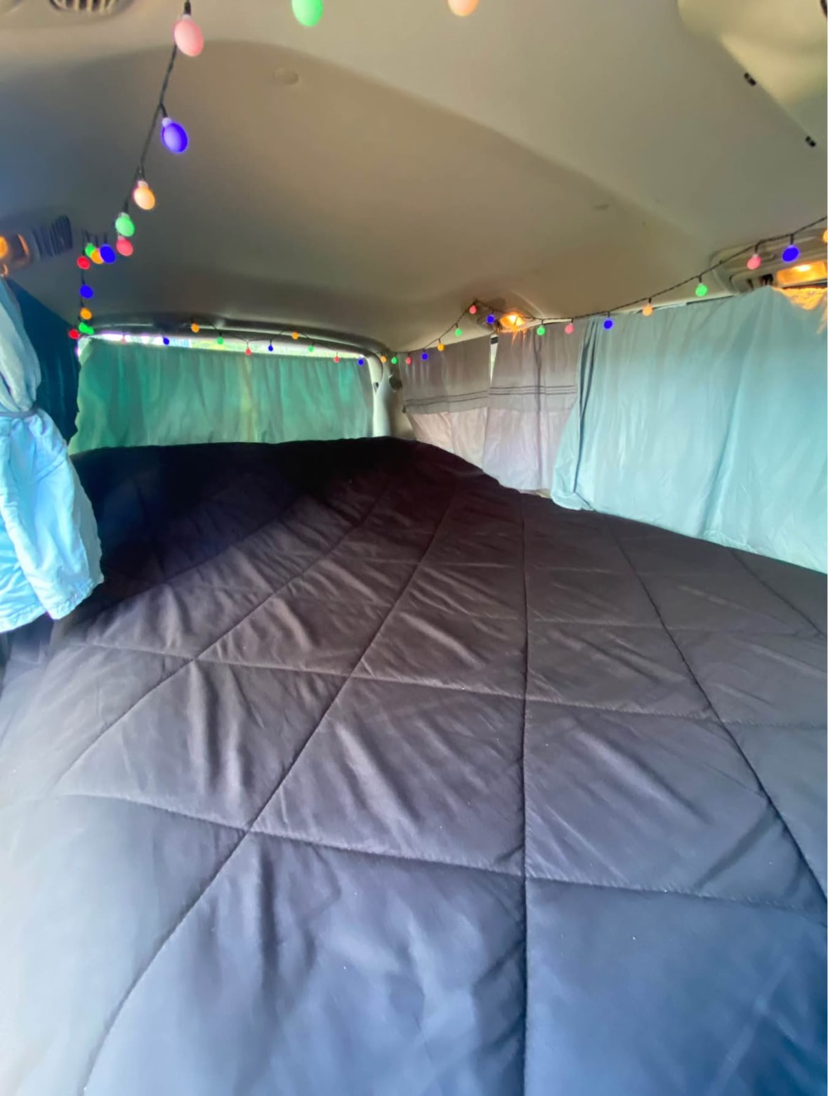 Cozy RV rental interior with a dark quilted bed, blue curtains, and colorful string lights for a comfortable road trip.. Toyota HIACE 2004