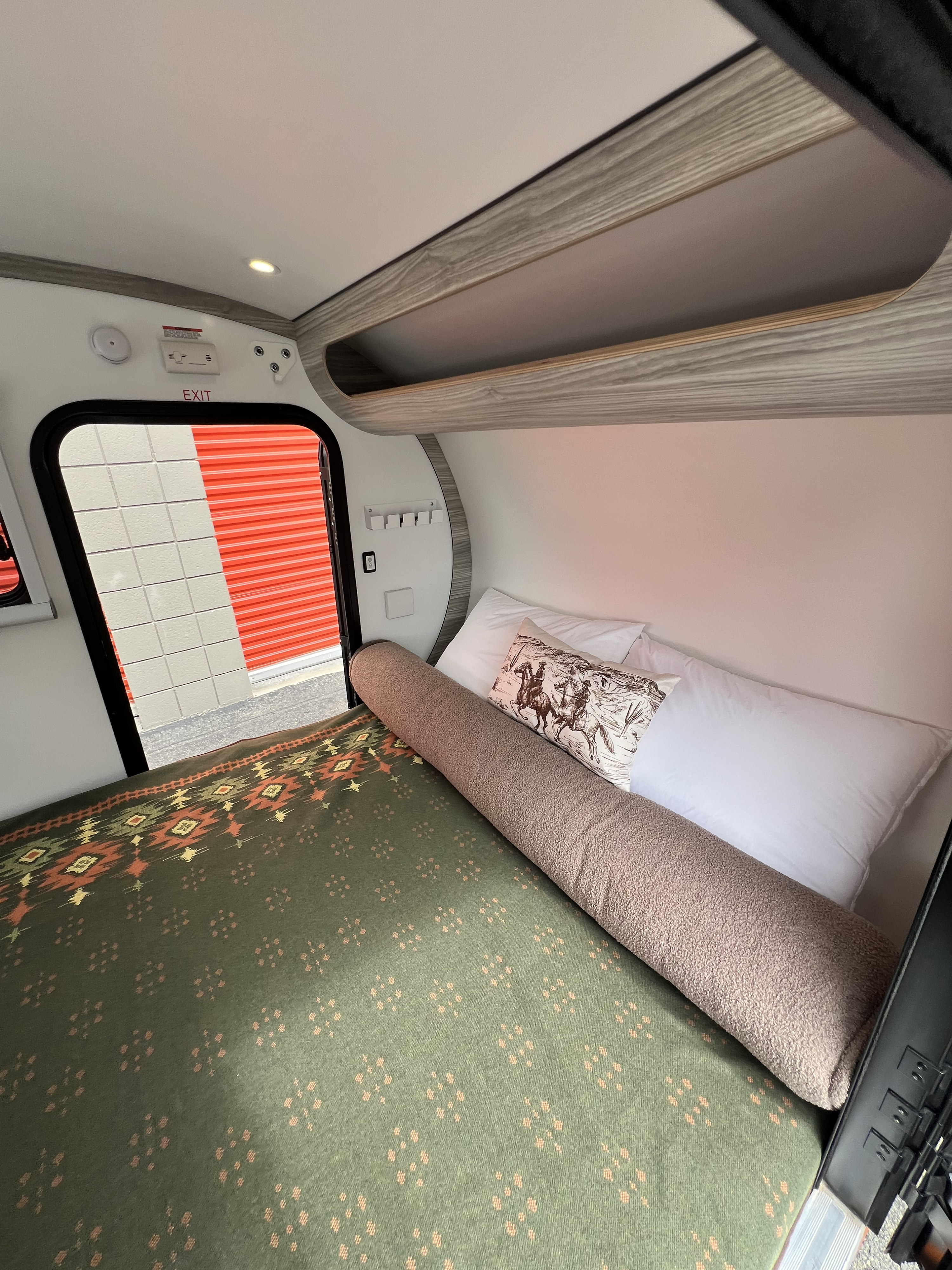 This cozy RV rental features a bed with patterned blanket, pillows, and overhead storage.. Bean Trailer Bean Stock 2.0 2024