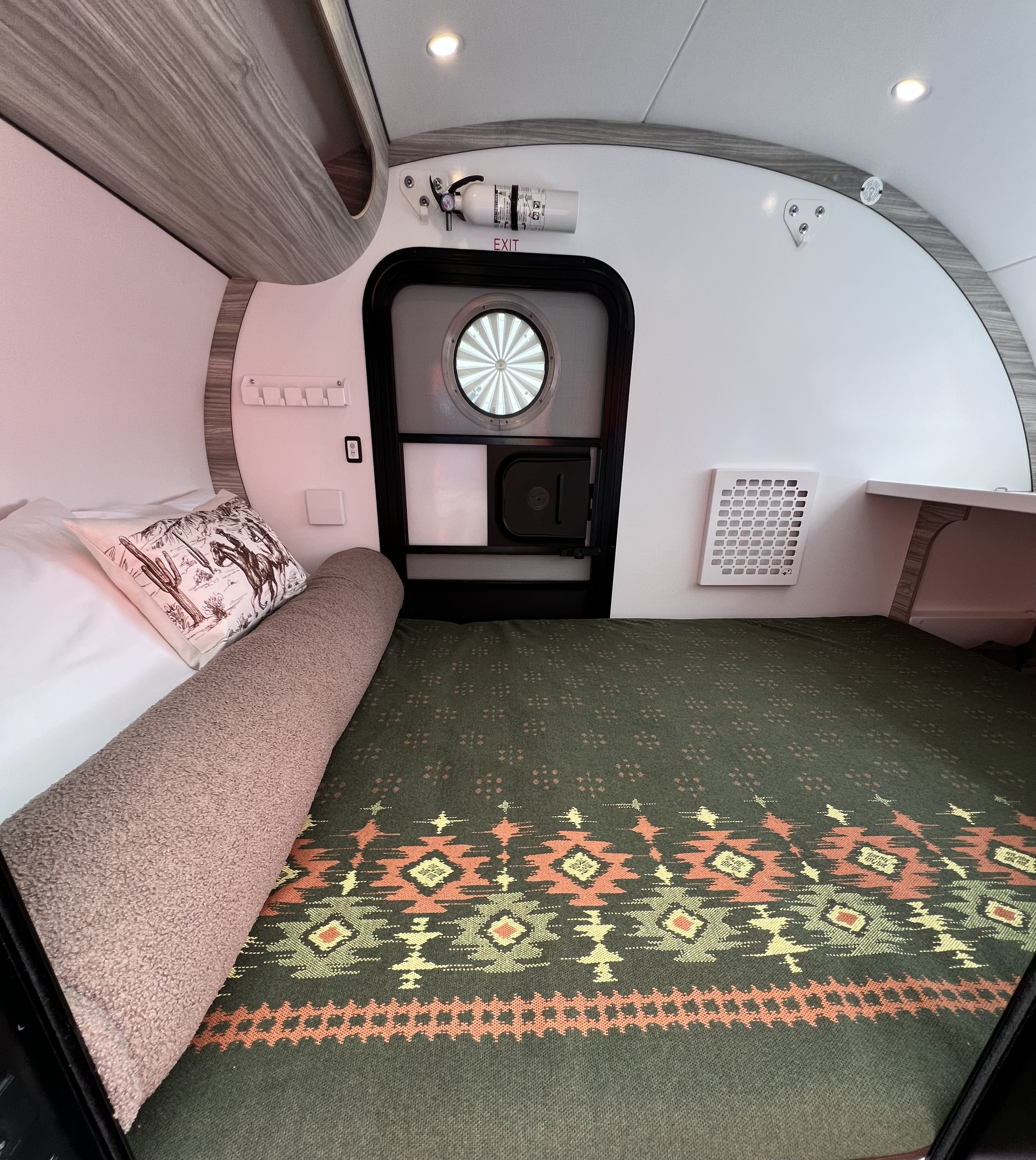 Cozy RV rental interior showcasing a bed with a vibrant patterned blanket, exit door, and fire extinguisher.. Bean Trailer Bean Stock 2.0 2024