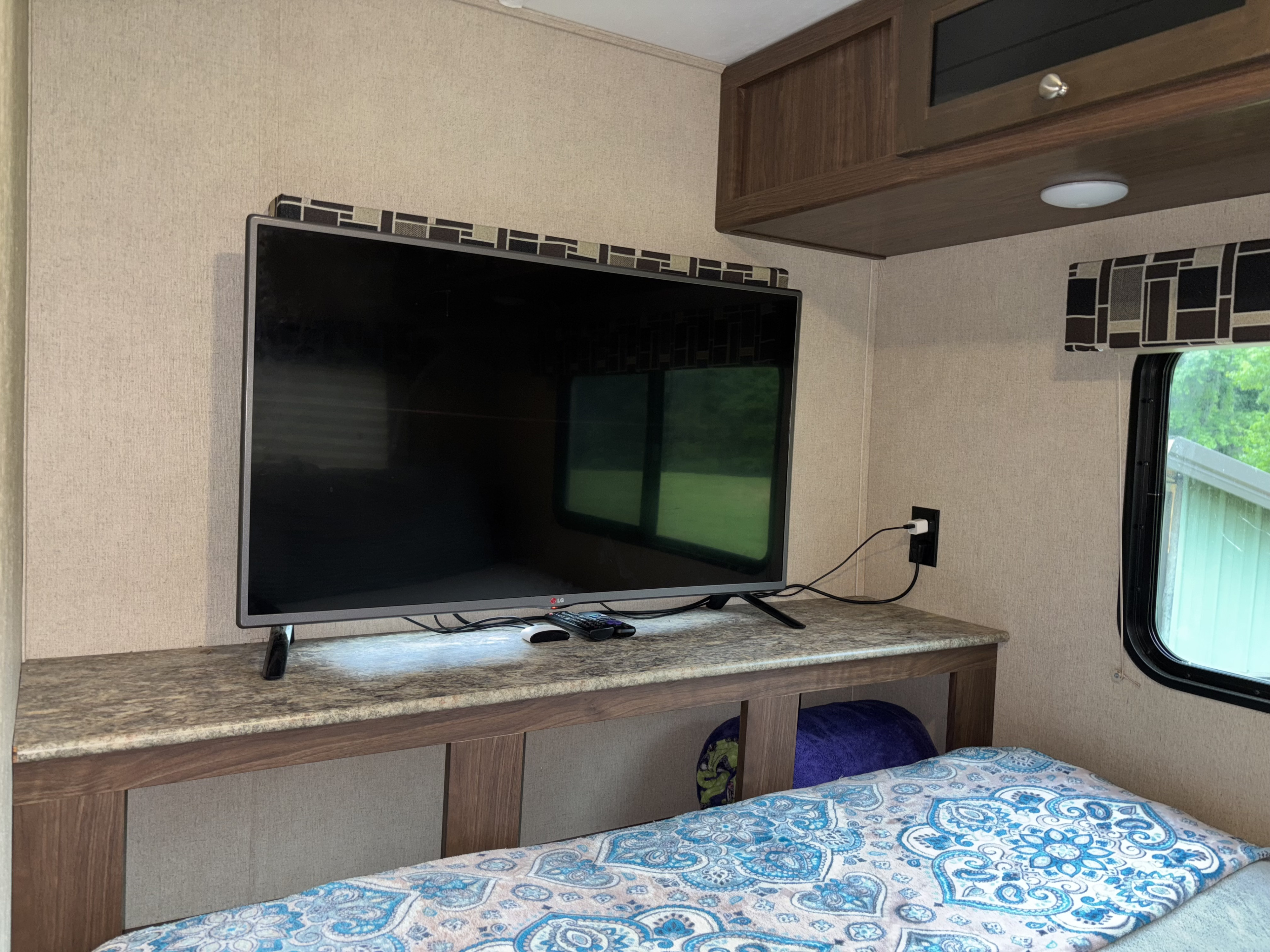Inside a cozy RV rental: a flat-screen TV on a stone-look countertop, overhead cabinets, and a bed with a blue patterned blanket.. Dutchmen Coleman Light LX 2018