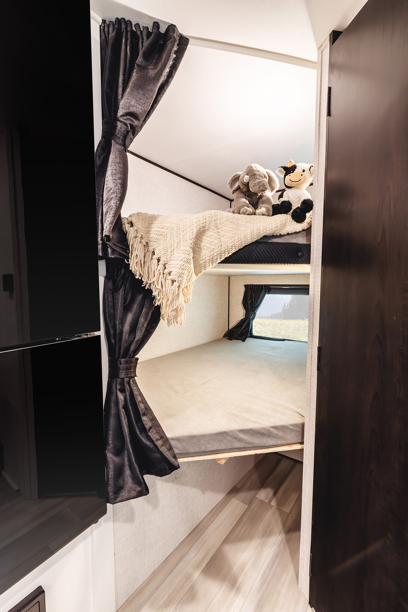 Inside an RV rental, two bunks with dark curtains. Stuffed animals rest on the top bunk, with a scenic window below.. Go Play 188BHS 2025