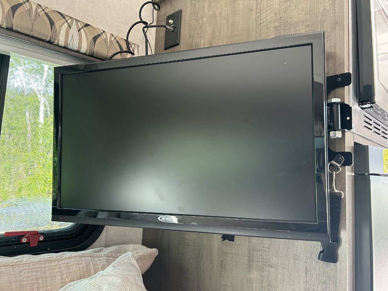 A black Jensen TV is wall-mounted and secured with a strap inside an RV rental. Pillows and a window are also visible.. Forest River R-Pod 172 2019