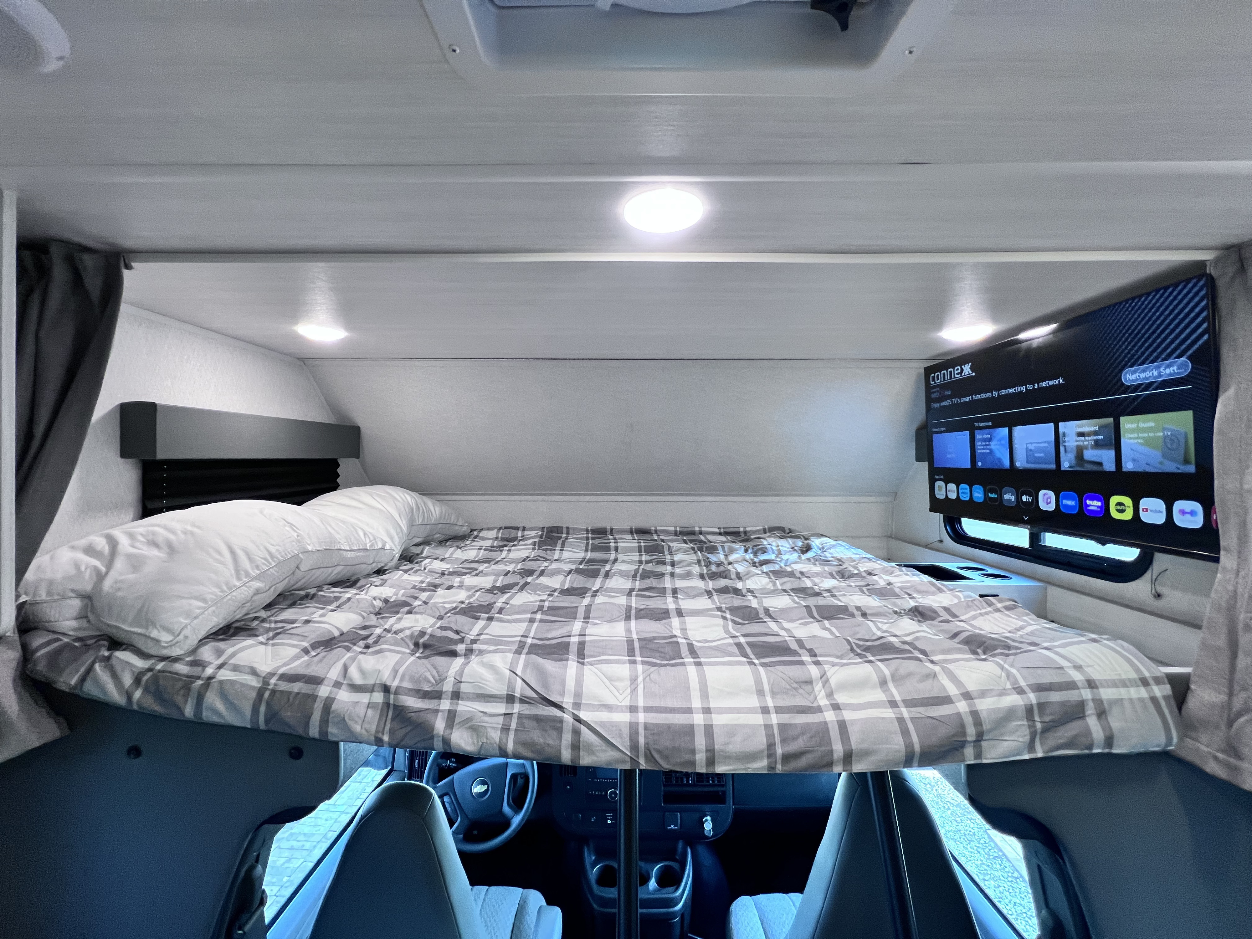 Comfortable overhead bed in an RV rental, featuring a smart TV and cozy plaid bedding above the driver's cabin.. East to West Entrada 210NSC 2025