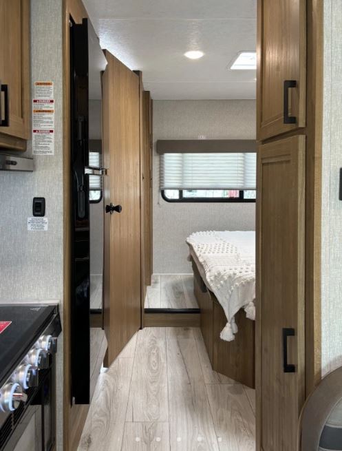 Inside a cozy rv rental: compact kitchen area, narrow hall, and a bright bedroom with a window. Features light wood flooring.. Coachmen Leprechaun 2025