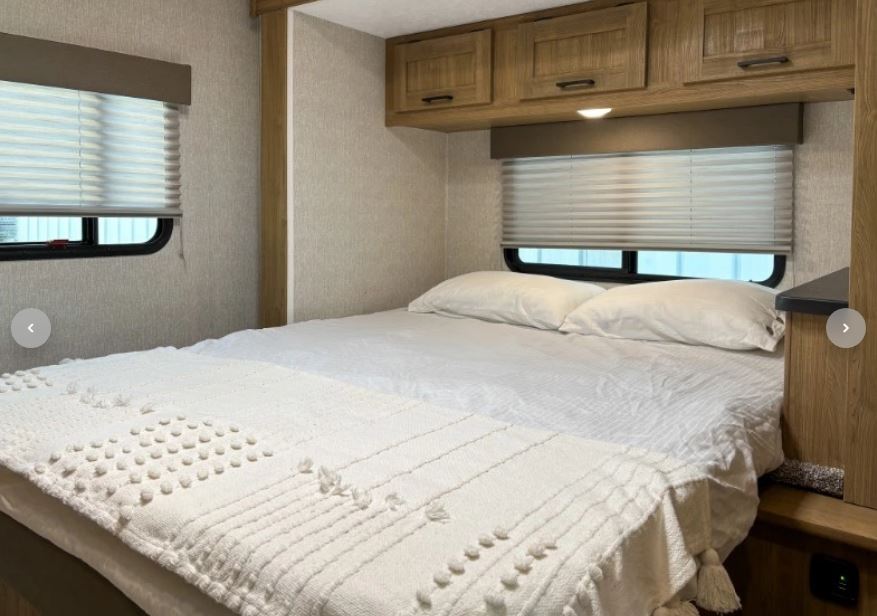 Cozy RV rental bedroom with white bedding, window blinds, and overhead cabinets, offering comfort for your travel adventures.. Coachmen Leprechaun 2025
