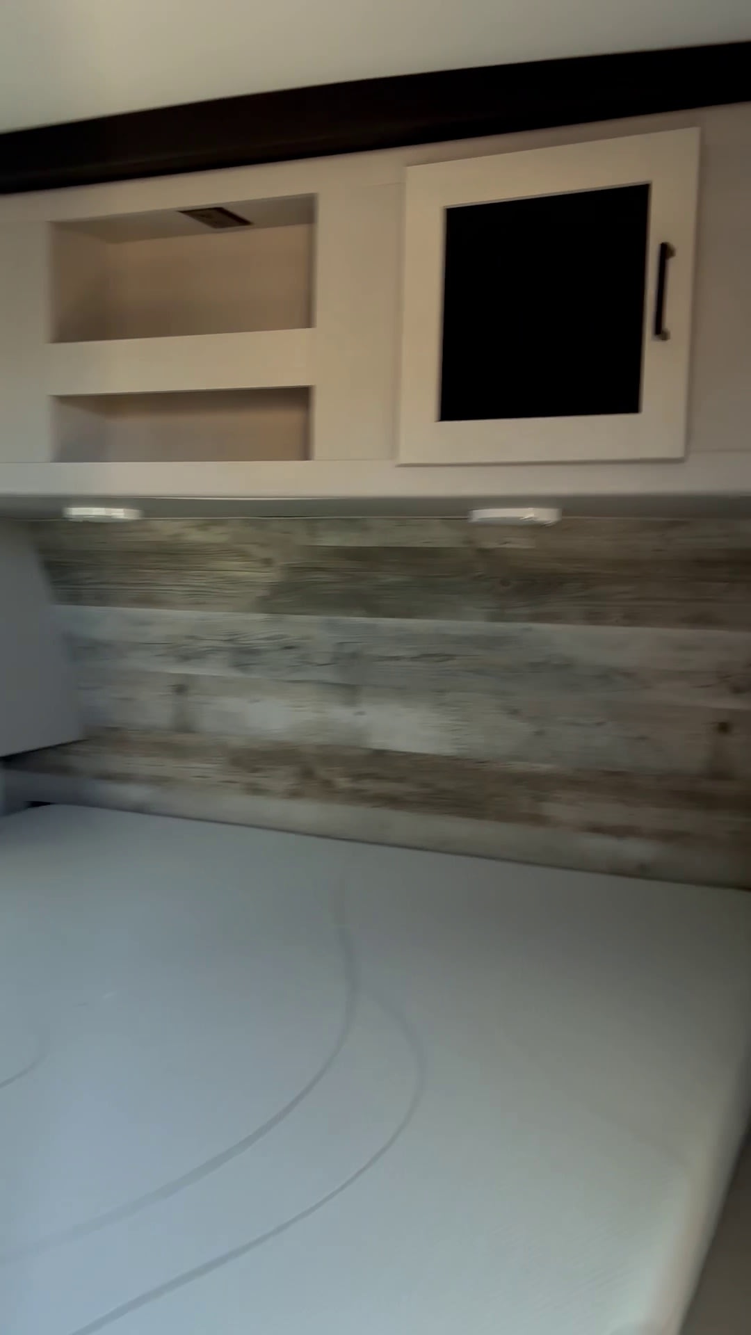 An RV rental bedroom with a white mattress, rustic wood panel headboard, and practical overhead storage including open shelves and a cabinet.. Heartland RVs Mallard 2022