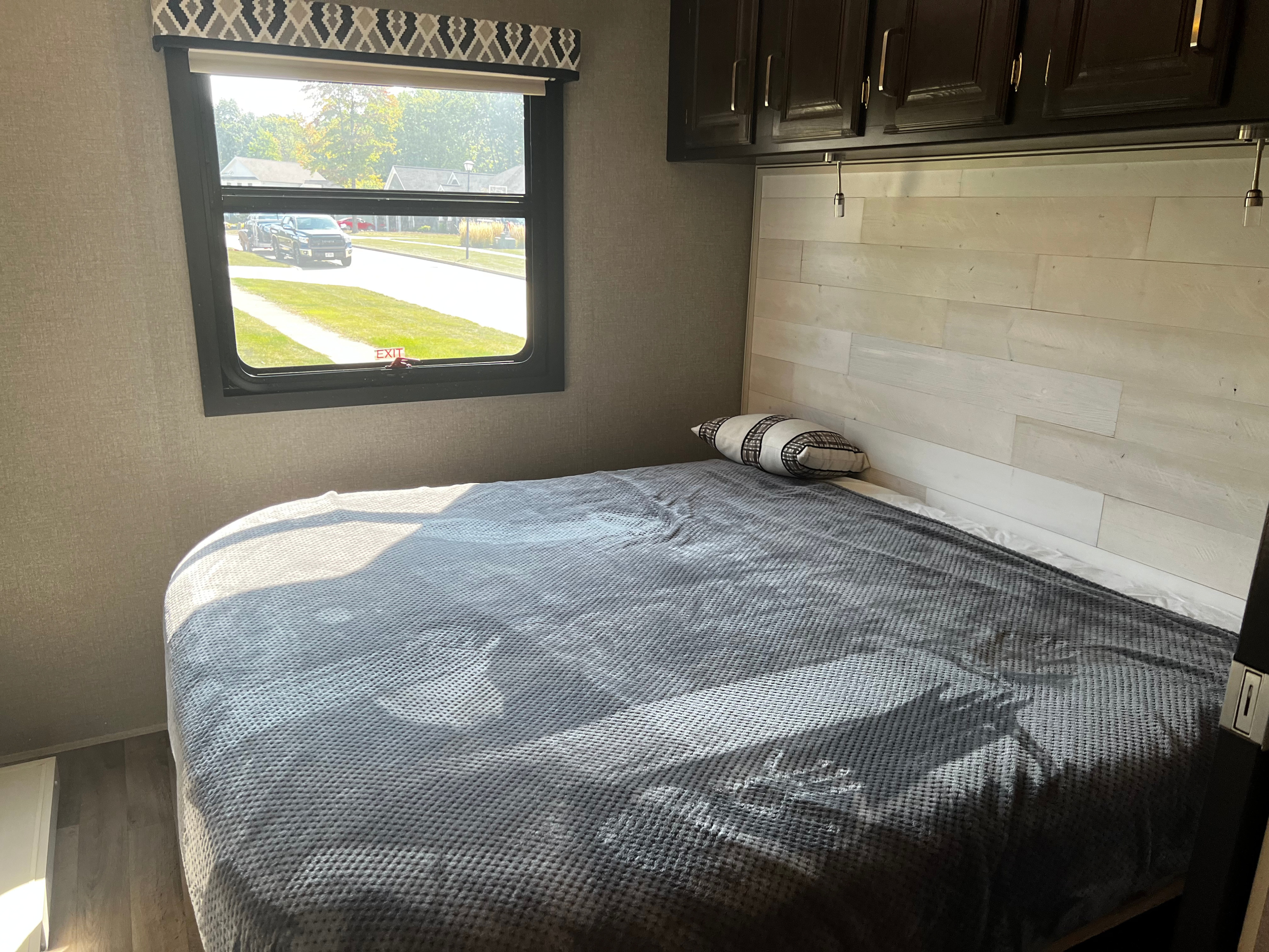 RV rental bedroom: a bed with a gray blanket, window overlooking a street, and dark wood overhead cabinets.. Jayco Precept 2020