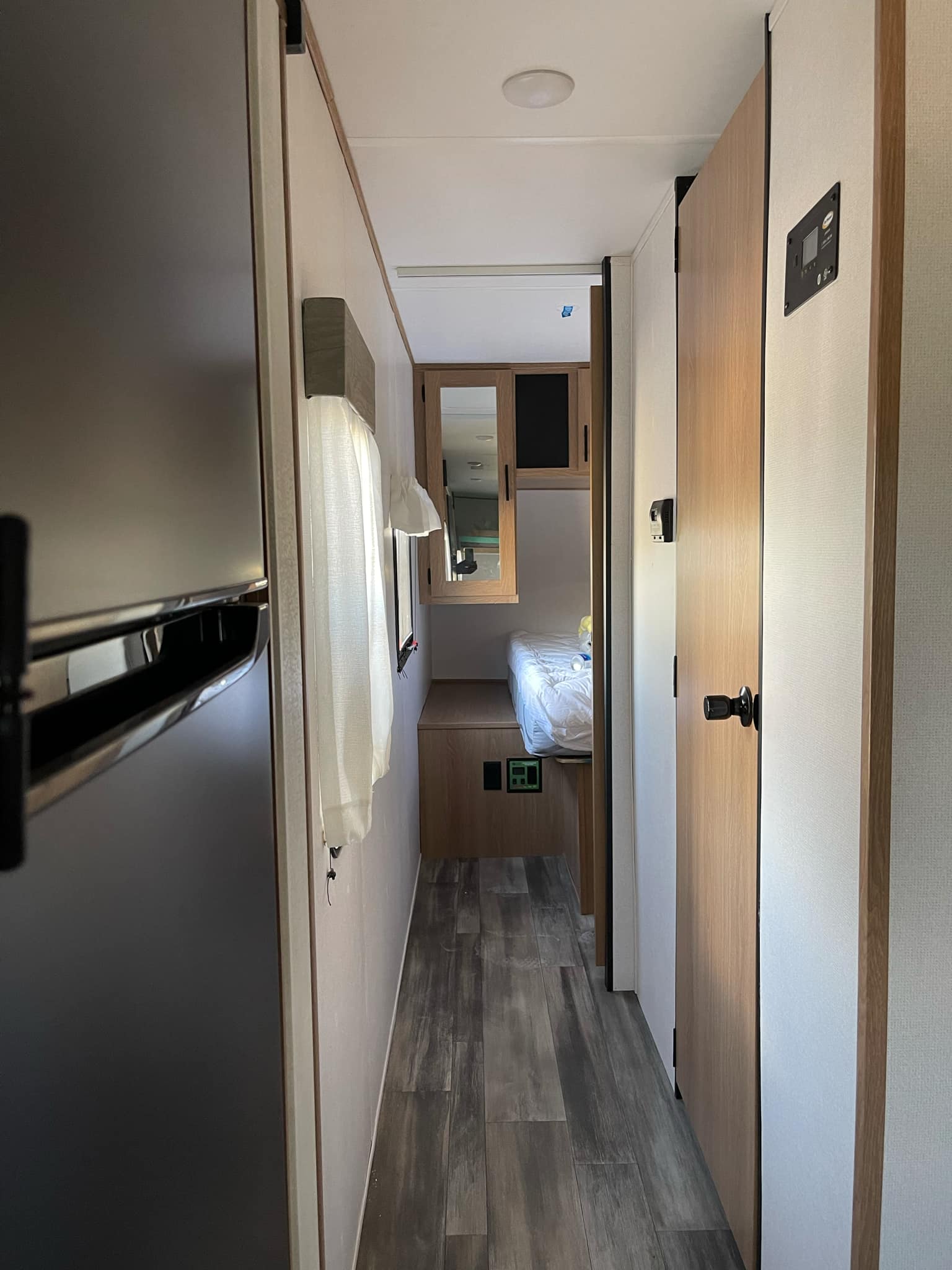 A hallway view inside an RV rental. A dark refrigerator is on the left, a control panel and wooden door on the right, leading to a bed.. Avenger Avenger Trailer 2022