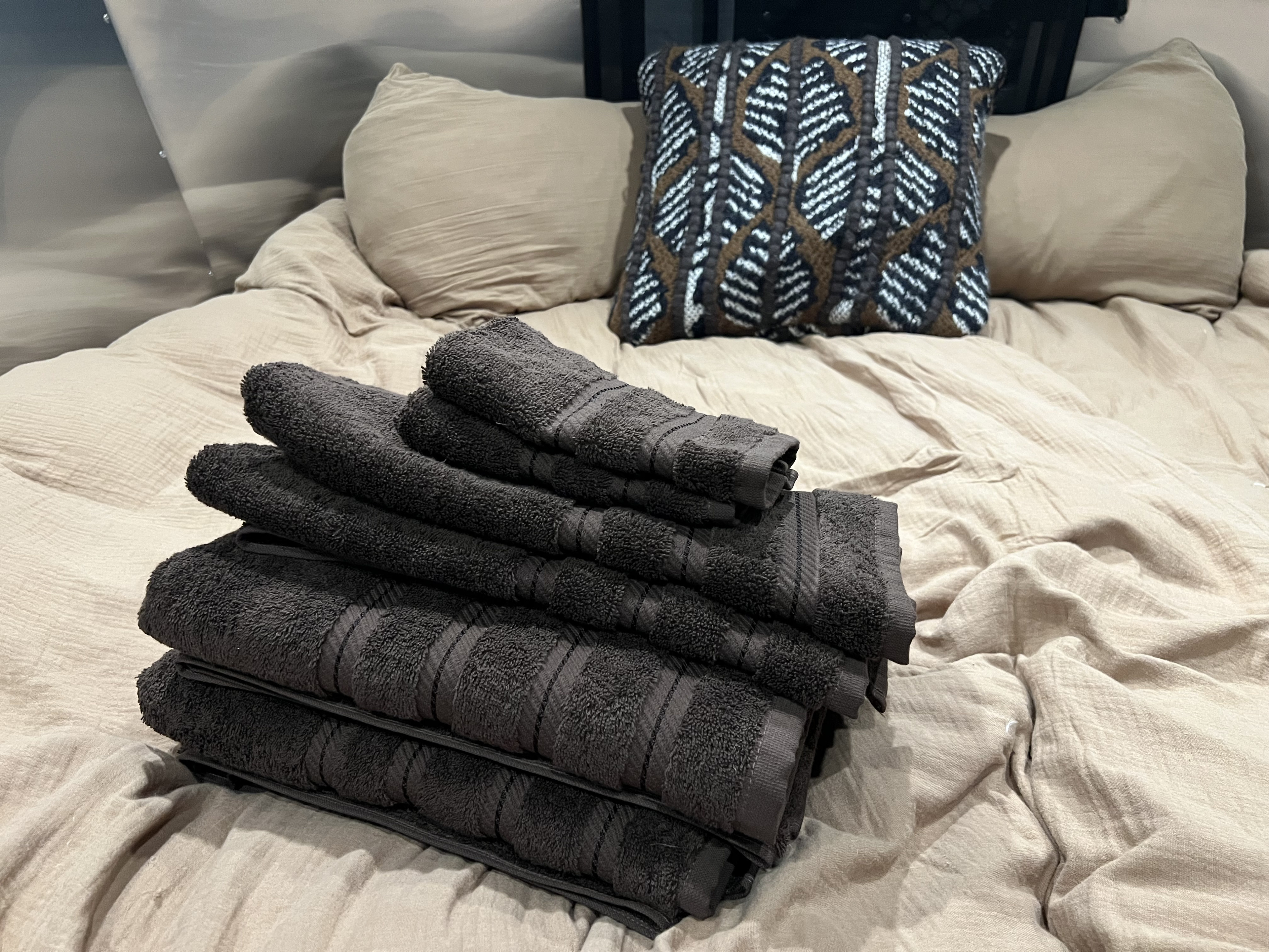 Plush dark towels stacked on a cozy bed with neutral and patterned pillows inside an RV rental, ready for guests.. Airstream Basecamp 2023
