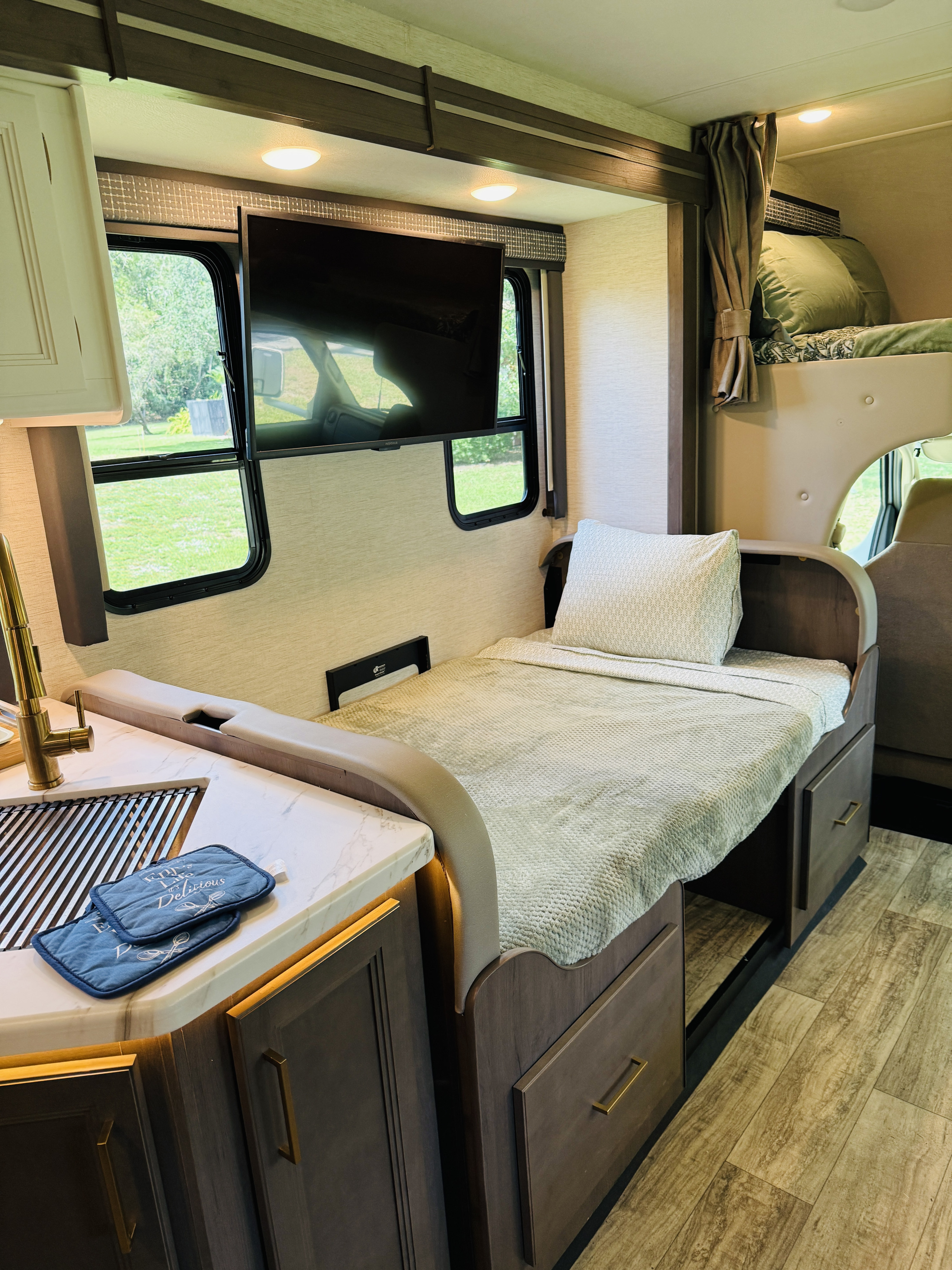 Bright RV rental interior: kitchen with sink & gold faucet, cozy bed, upper bunk, and wall-mounted TV. Ready for travel!. Thor Chateau 2024