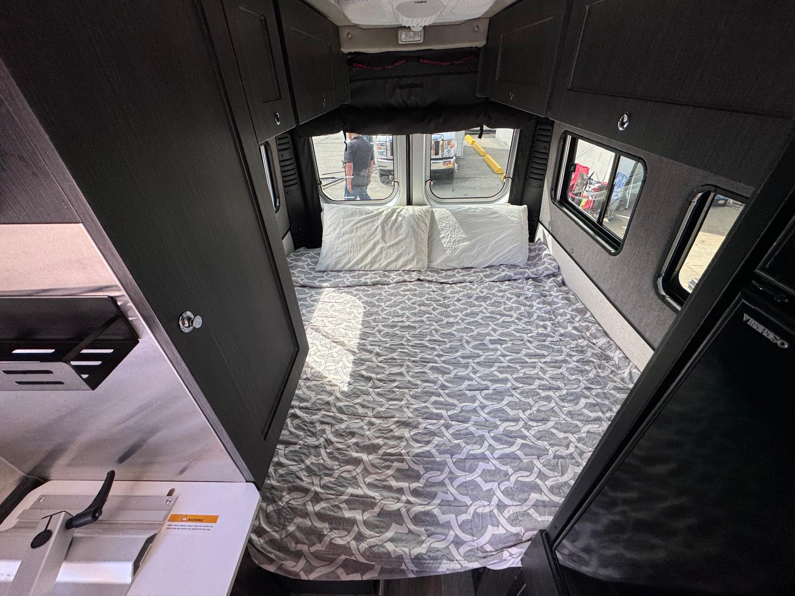 RV rental camper van's cozy sleeping area: a gray patterned bed, dark storage cabinets, and outside views through windows.. Roadtrek Chase Class B 2022