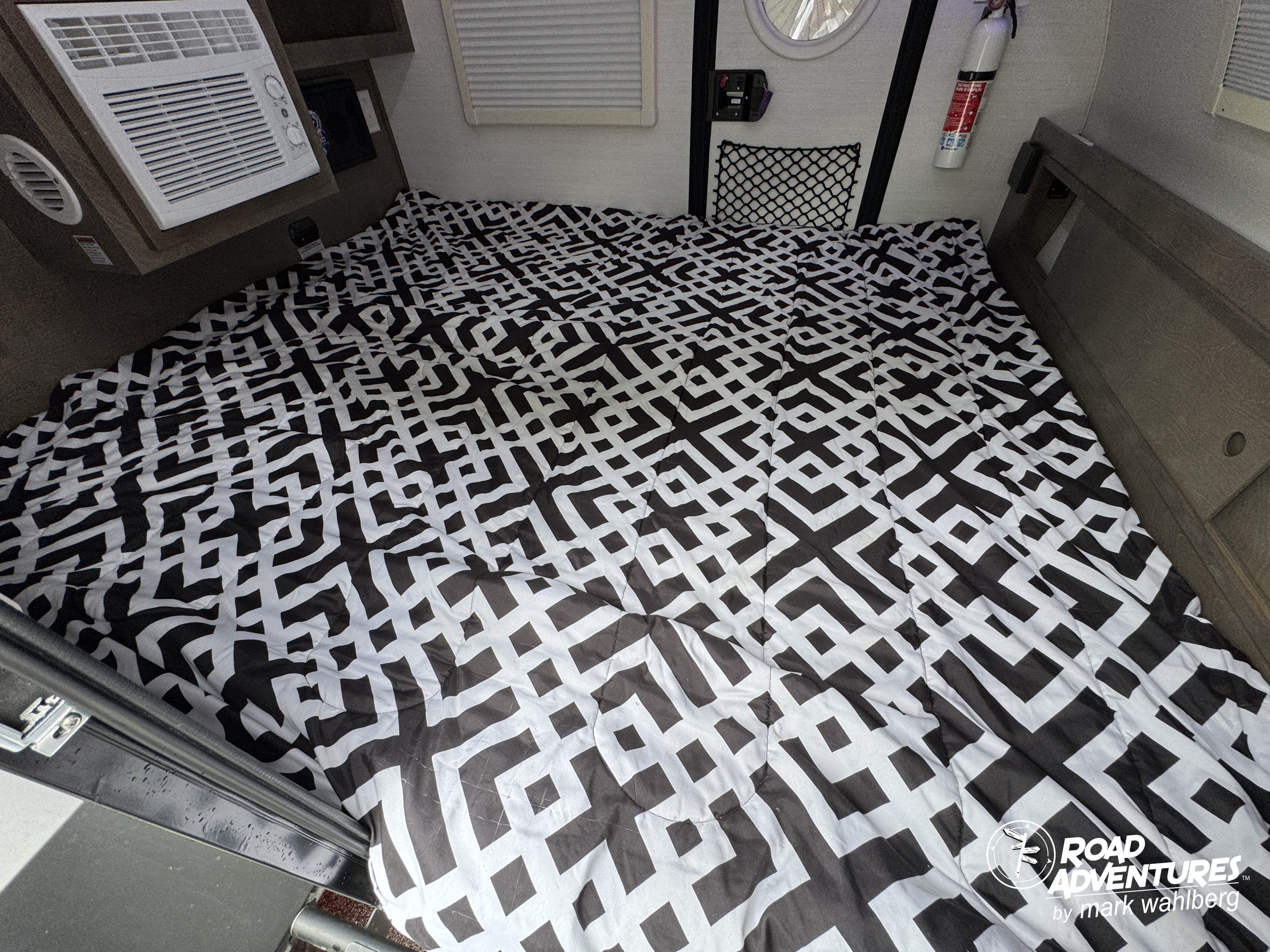 RV rental interior: A compact bedroom with a black and white geometric patterned bed. An AC unit is on the left; a window is above.. nuCamp T@G XL 2025