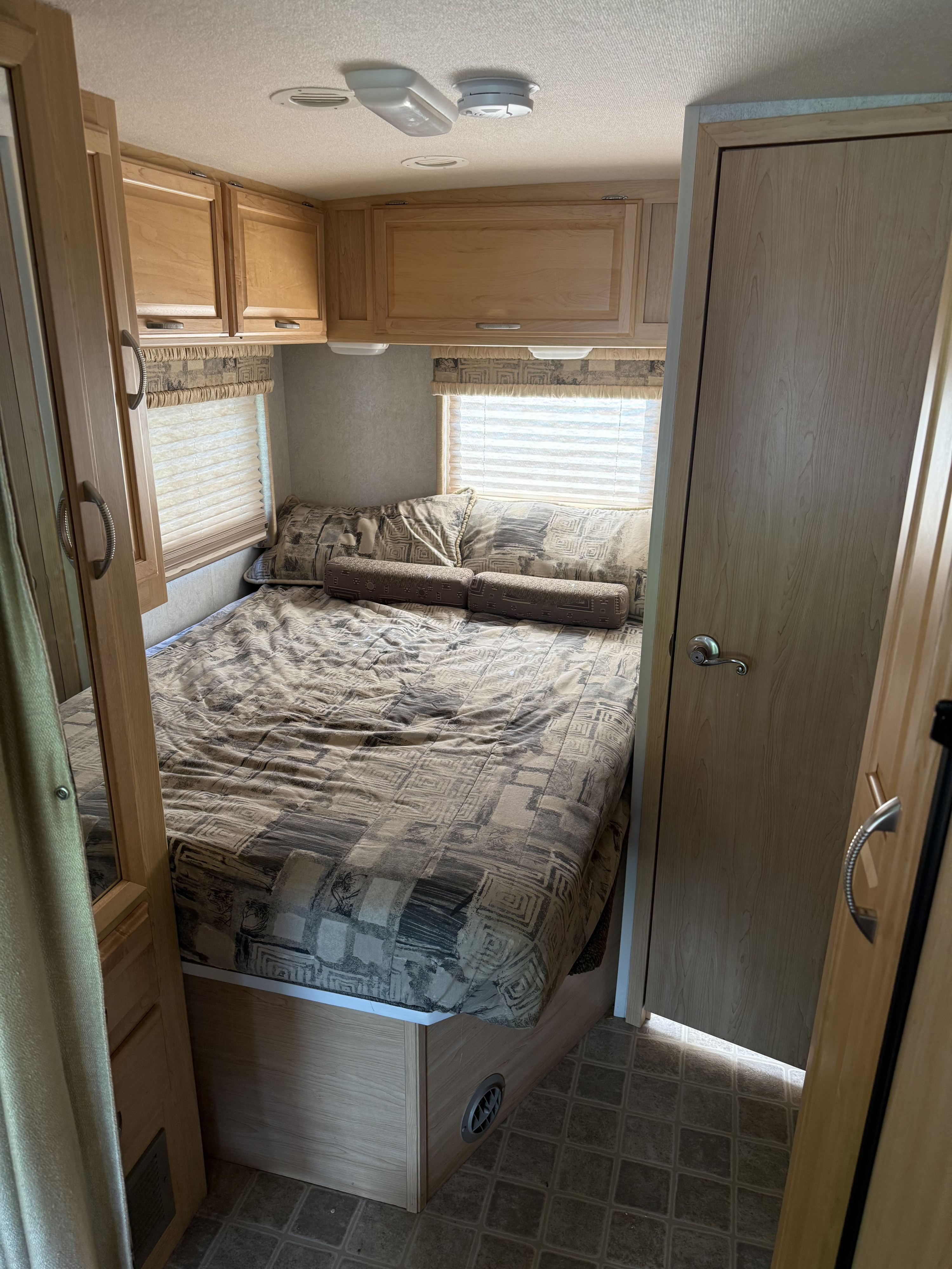 This RV rental bedroom shows a patterned bed, wooden overhead cabinets, a window with blinds, and tiled flooring.. Winnebago Aspect 2005