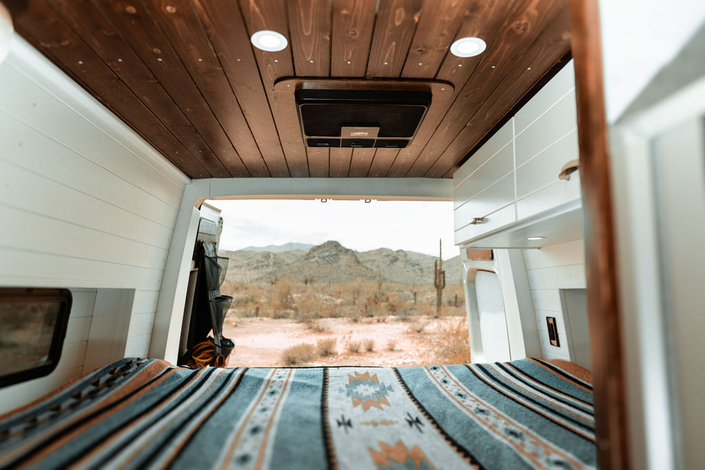 Cozy rv rental bed offers a desert view of mountains & cacti. Features wood ceiling, white paneled walls, & an AC unit.. Tommy Vans Mercedes Benz Sprinter 2024