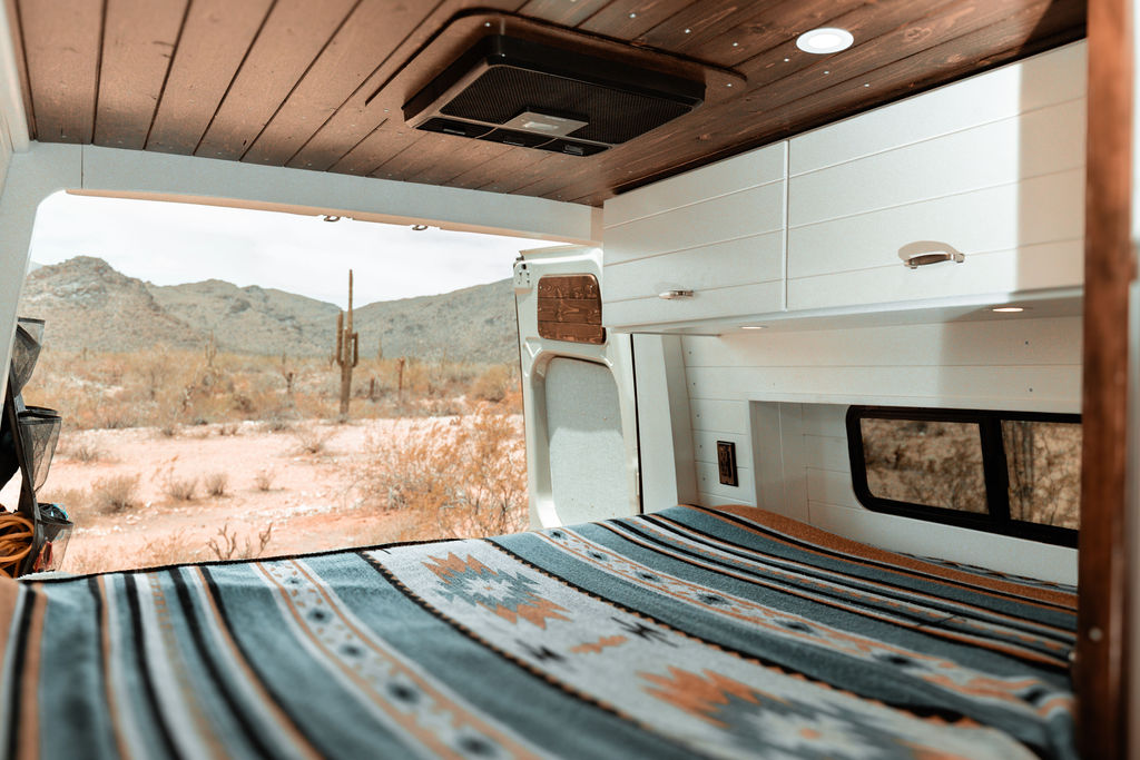 Inside a custom camper van rv rental: a cozy bed with a patterned blanket, looking out to a stunning desert landscape.. Tommy Vans Mercedes Benz Sprinter 2024