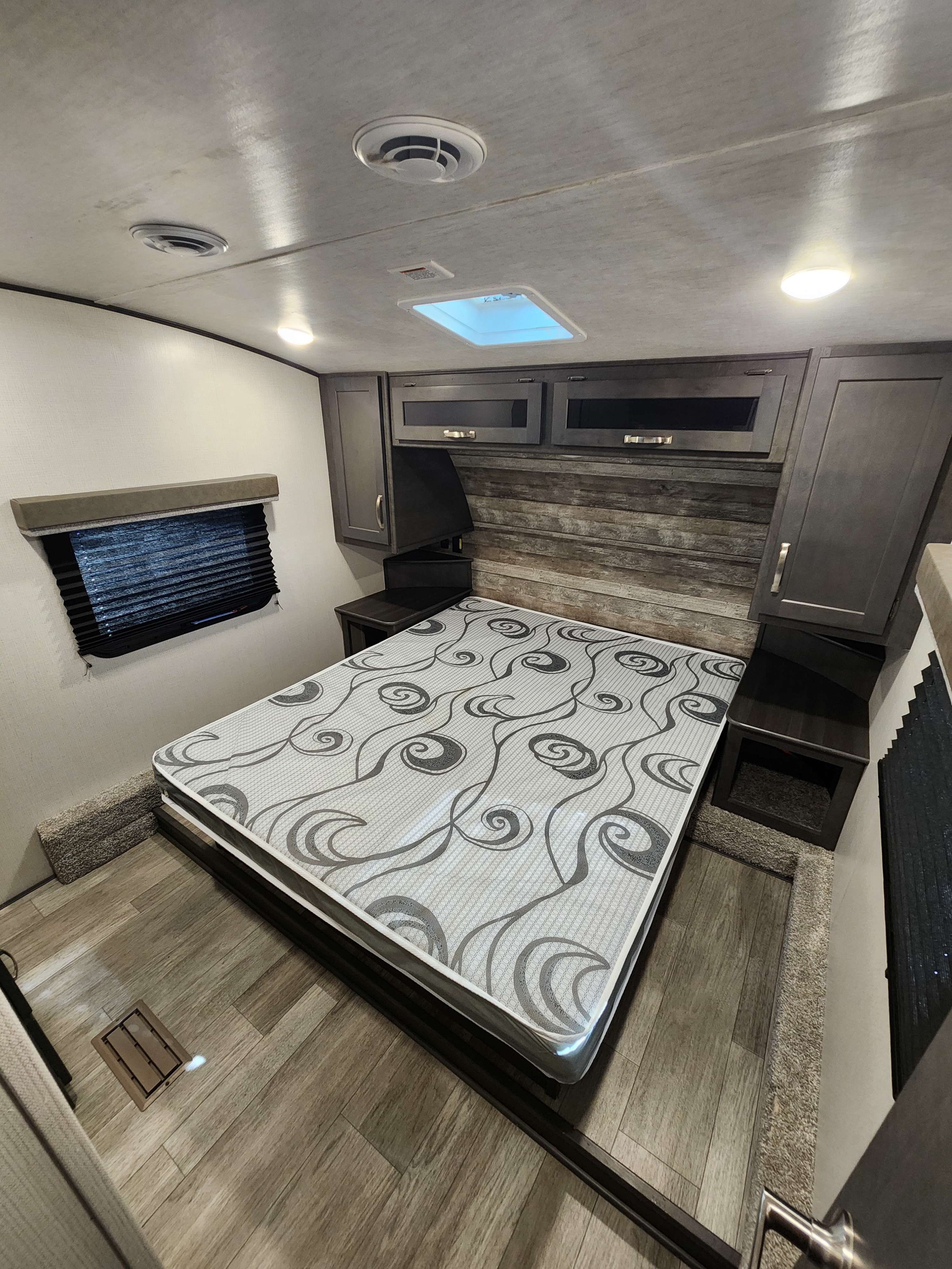Spacious RV rental bedroom features a comfy patterned bed, overhead storage, and modern wood-look flooring.. Keystone RV Cougar 2024