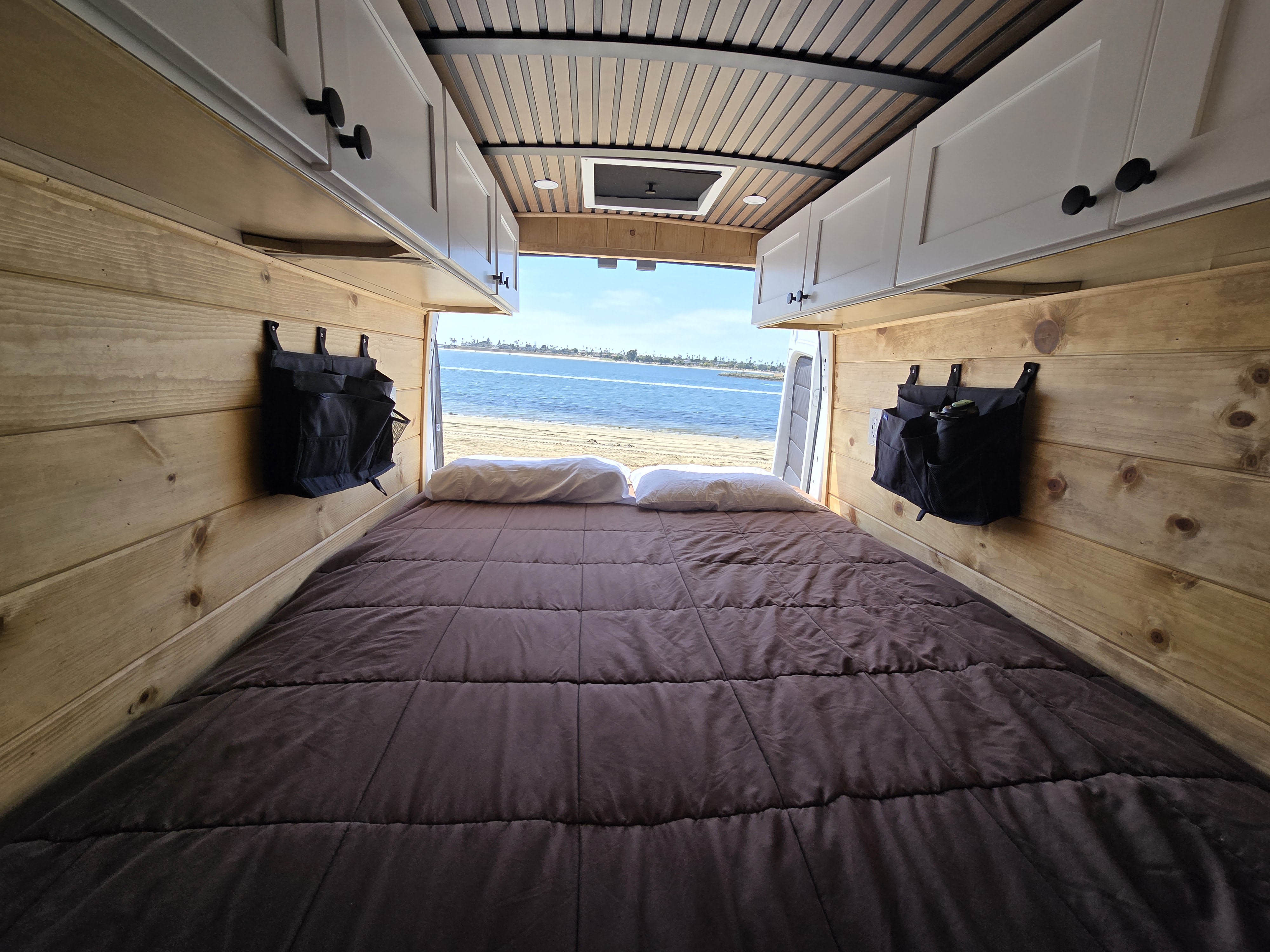 Cozy RV rental camper van interior: a bed with wood walls opens to a beautiful beach and ocean view.. Custom Ford Transit 350HD 2019
