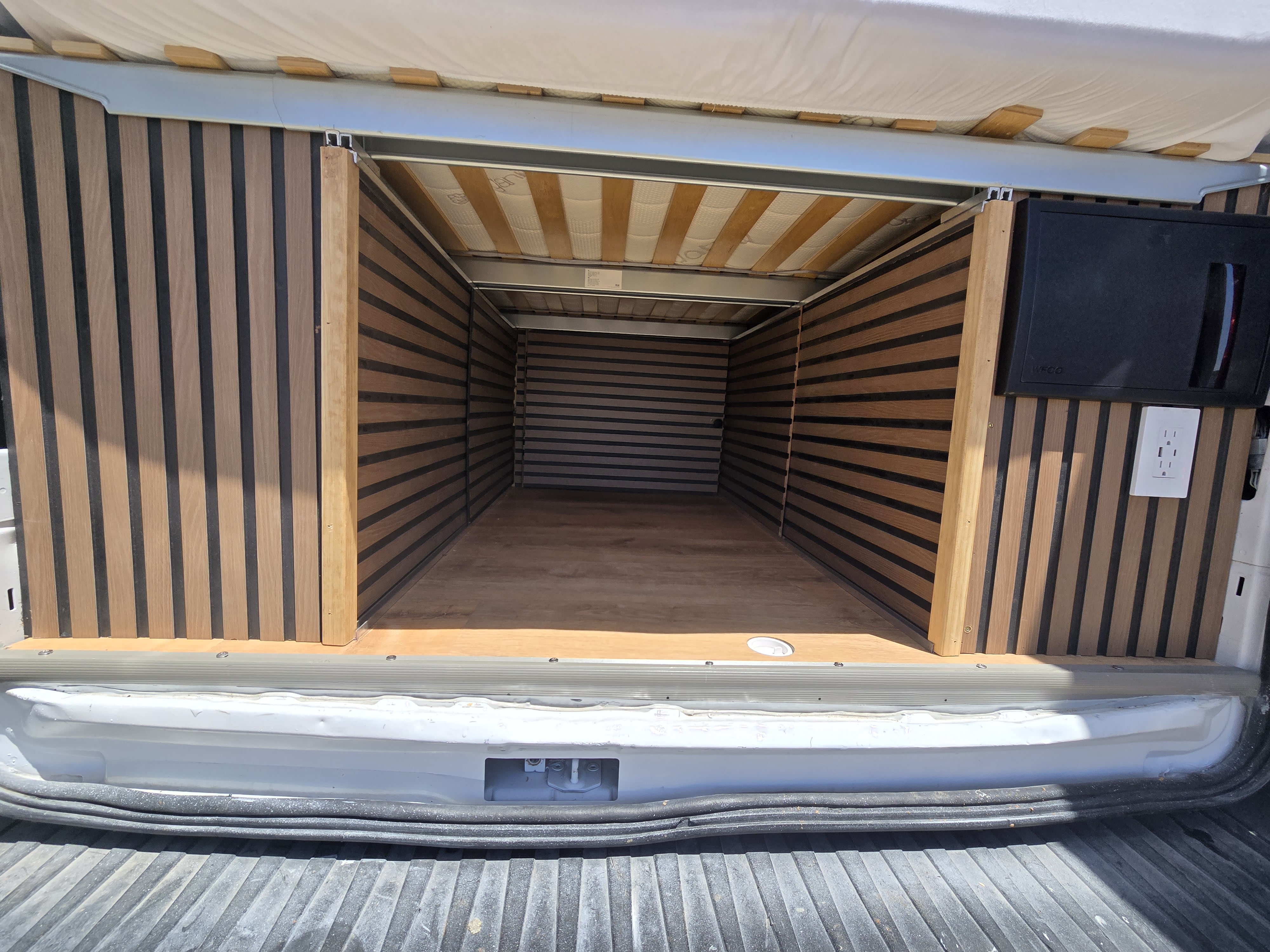 RV rental: Rear view of a camper van's wood-paneled interior, revealing storage space, a bed platform, & power outlets.. Custom Ford Transit 350HD 2019