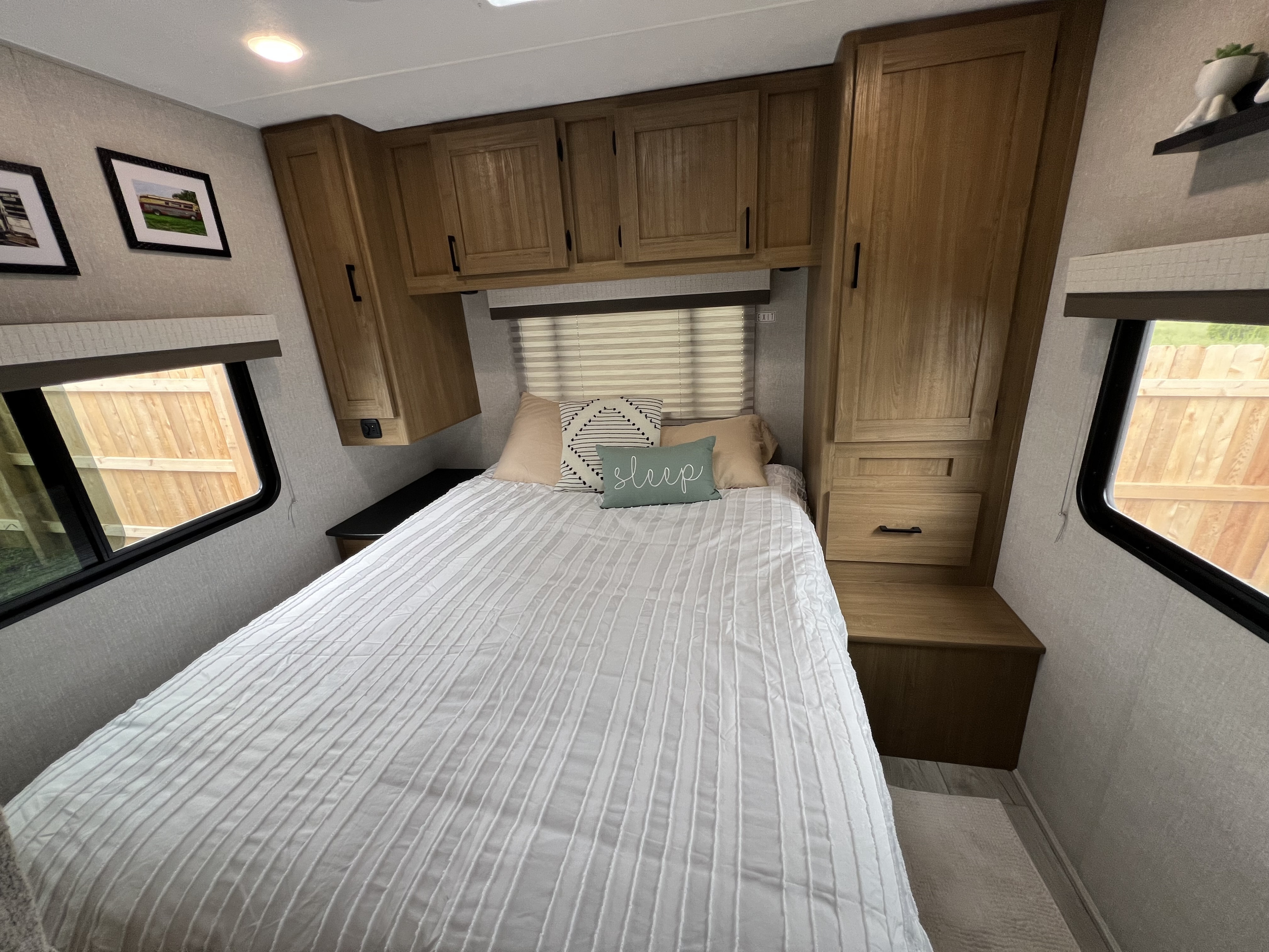 Cozy RV rental bedroom featuring a comfortable bed with white bedding, throw pillows, wooden cabinets, and bright windows.. Forest River Coachmen Leprechaun 2024