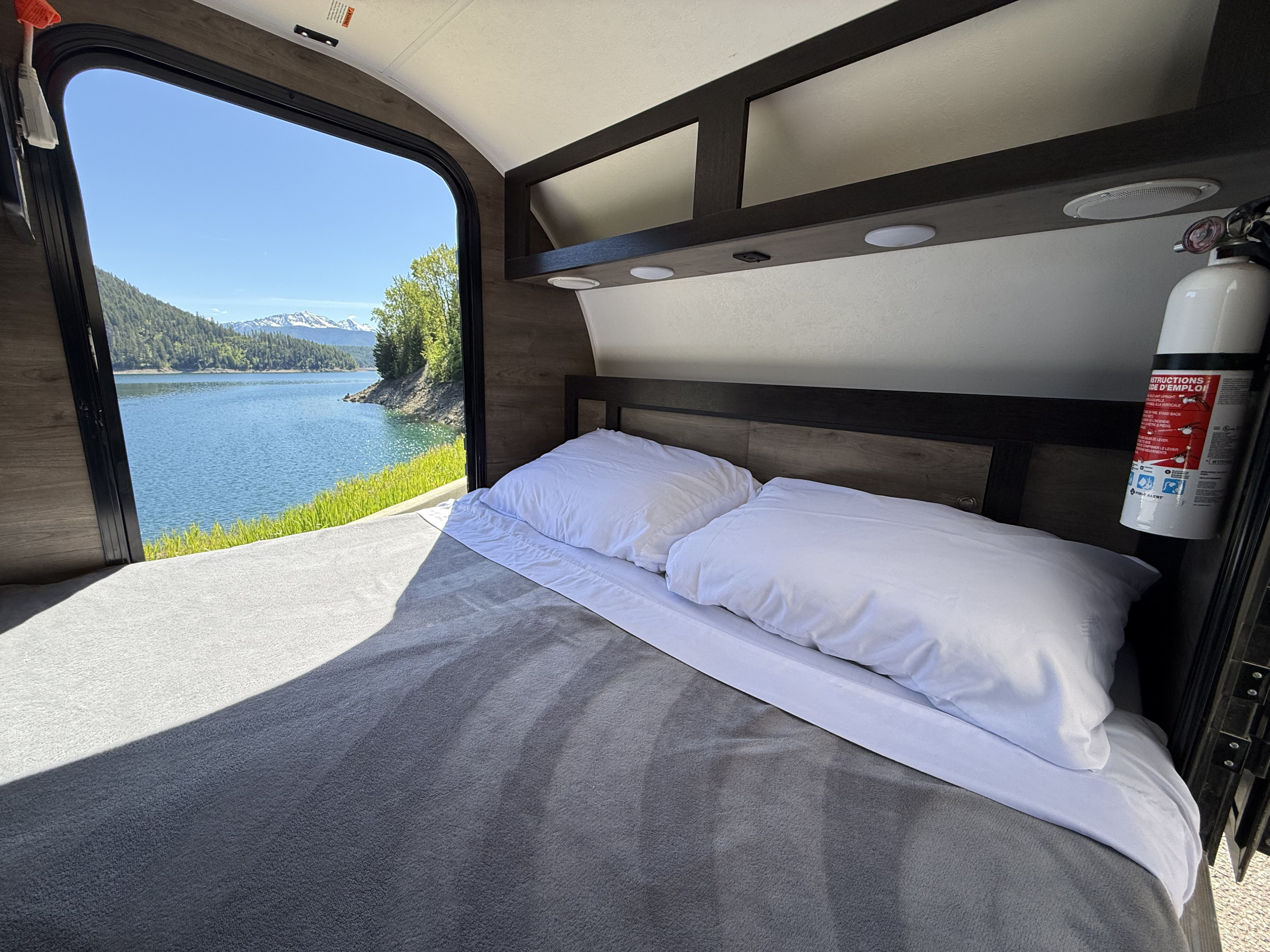 Inside an RV rental, a cozy bed faces a large window with a beautiful view of a lake and snow-capped mountains.. Braxton Creek Bushwhacker 2022