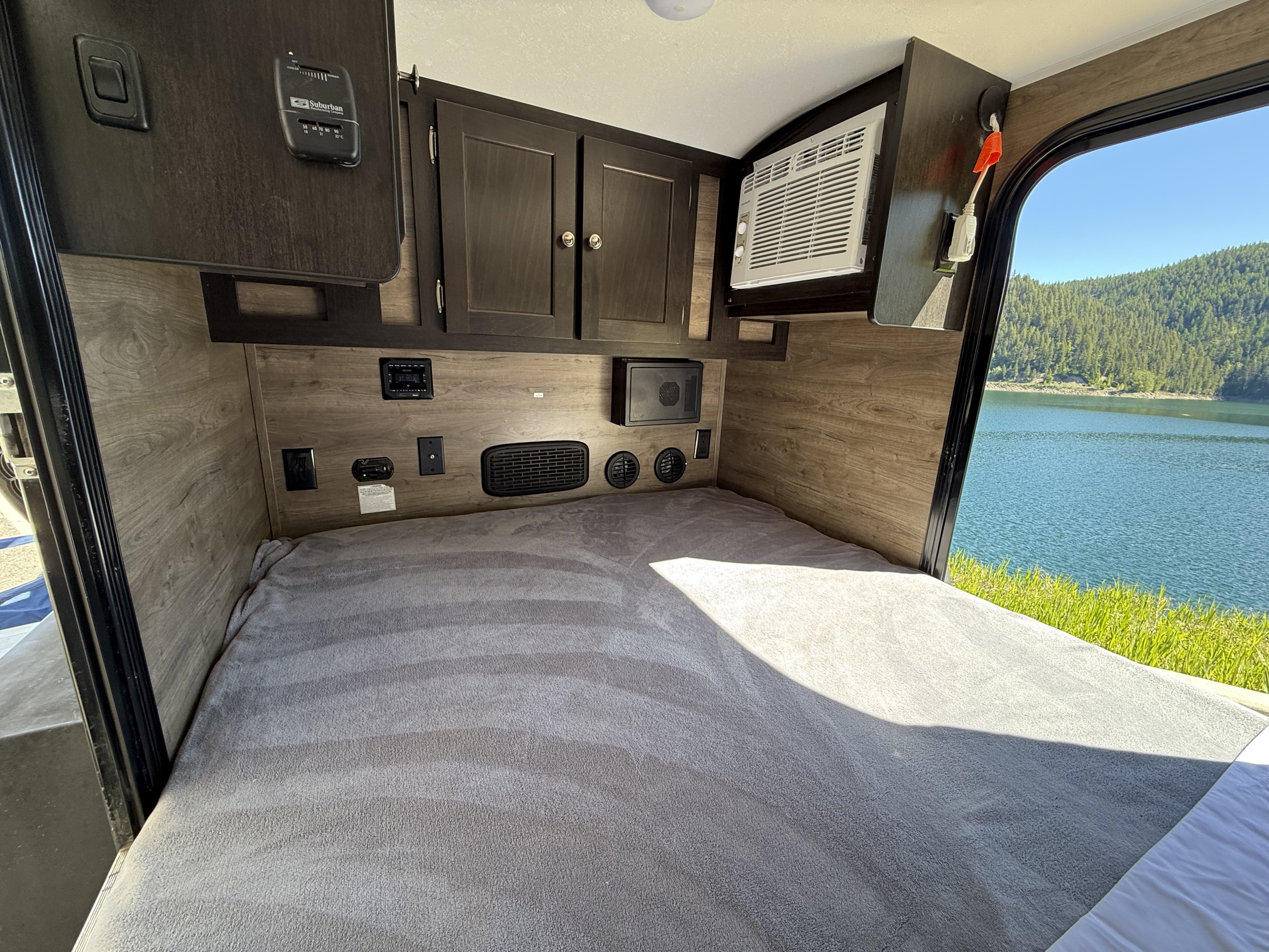 Inside a comfy RV rental: cozy bed, control panels, AC unit, & scenic lake view through the window overlooking mountains.. Braxton Creek Bushwhacker 2022