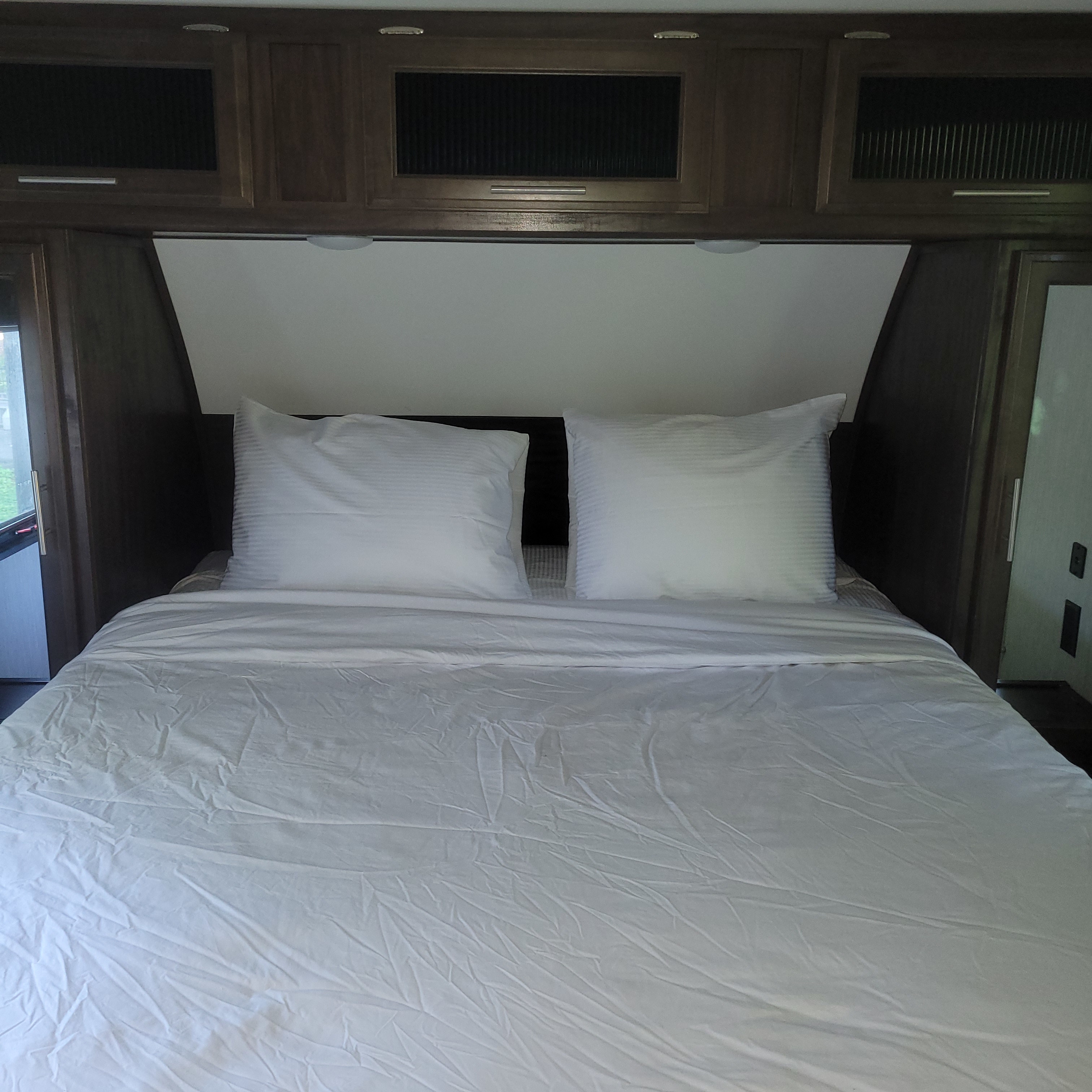 White sheeted bed in an RV rental, flanked by dark wood cabinets and a window, offering a cozy sleep space.. Forest River Cherokee Alpha Wolf 2020