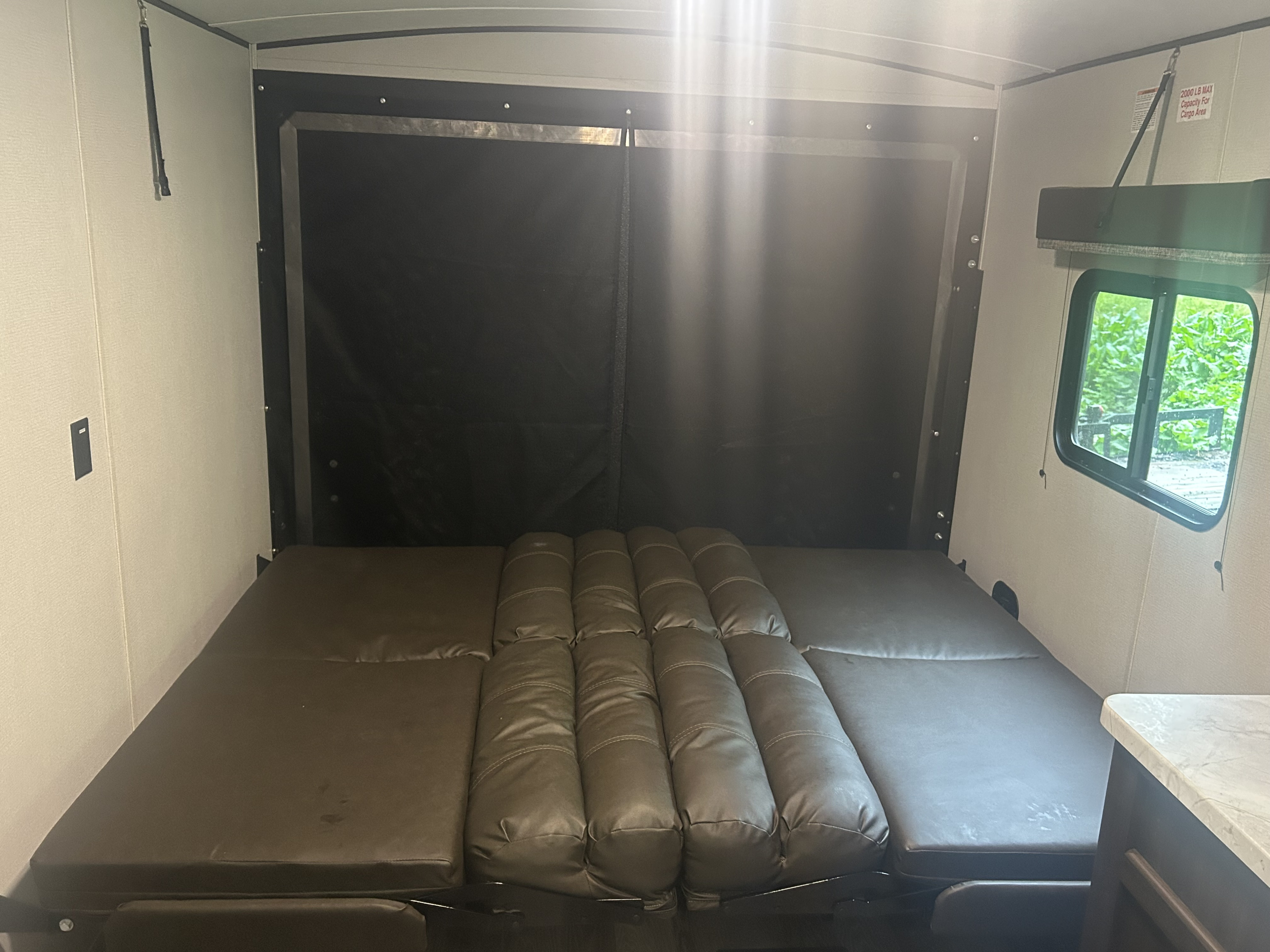 Spacious RV rental interior with a versatile brown convertible bed/sofa system, rear ramp door, and outdoor view.. Keystone Tailgator 2020
