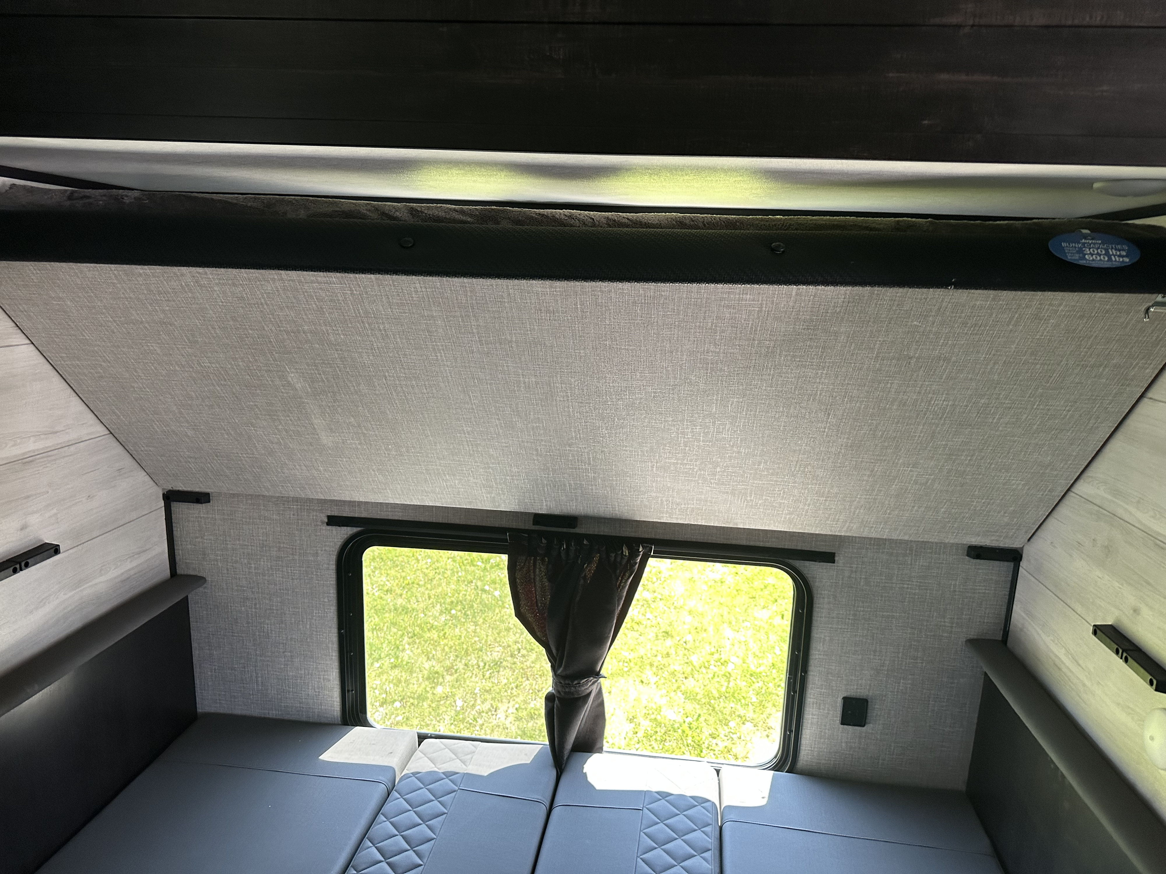 RV rental interior: A top bunk with capacity limits, above grey convertible seating, next to a window showing green grass outside.. Jayco Jay Flight 2023