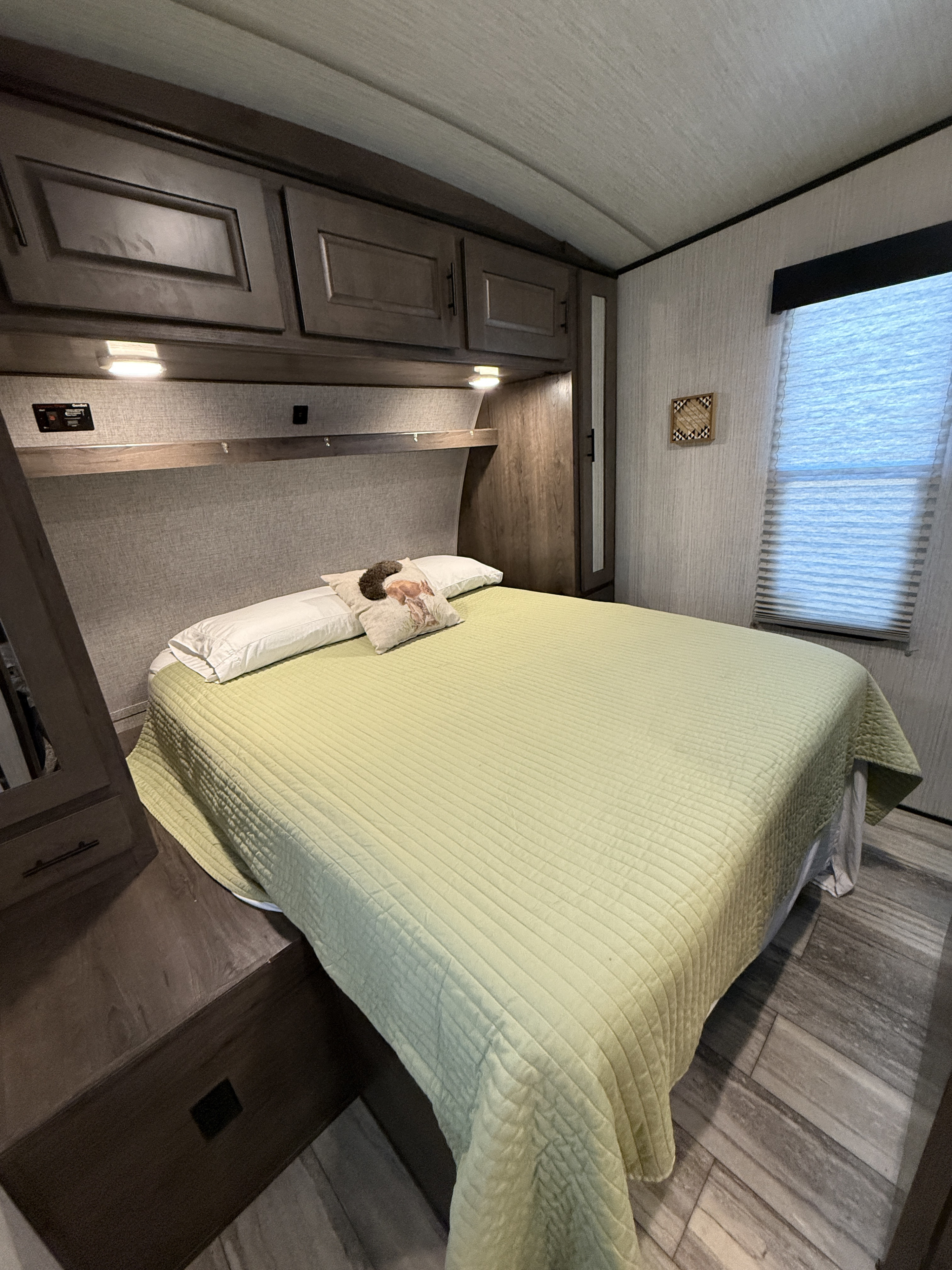 Cozy RV rental bedroom with a light green quilted bed, white pillows, dark wood cabinets, and a window with blinds.. Heartland Torque 2021
