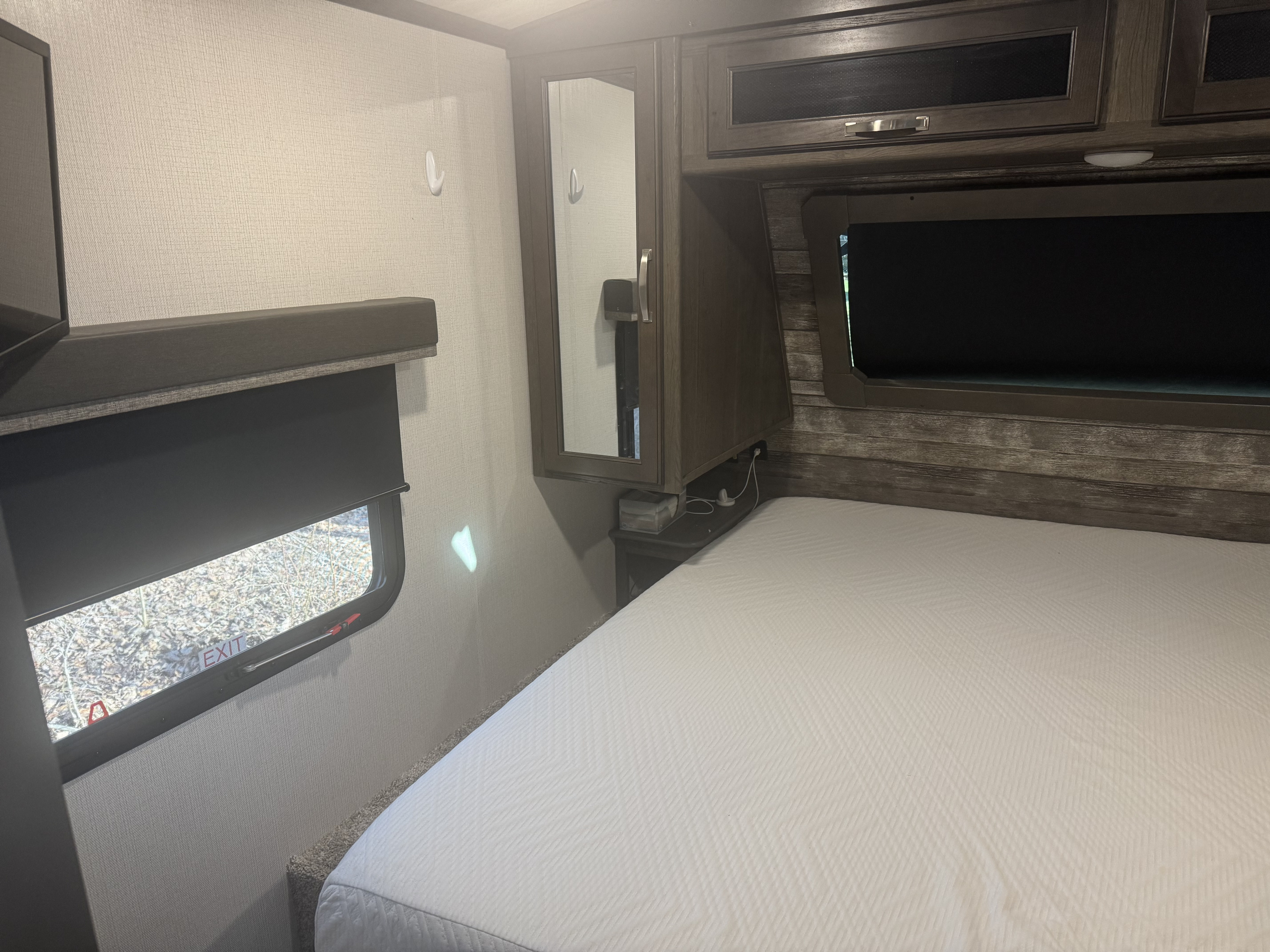 RV rental bedroom showcasing white bedding, dark wood cabinets, a bedside mirror, and window with emergency exit sign.. Keystone RV Cougar 2022