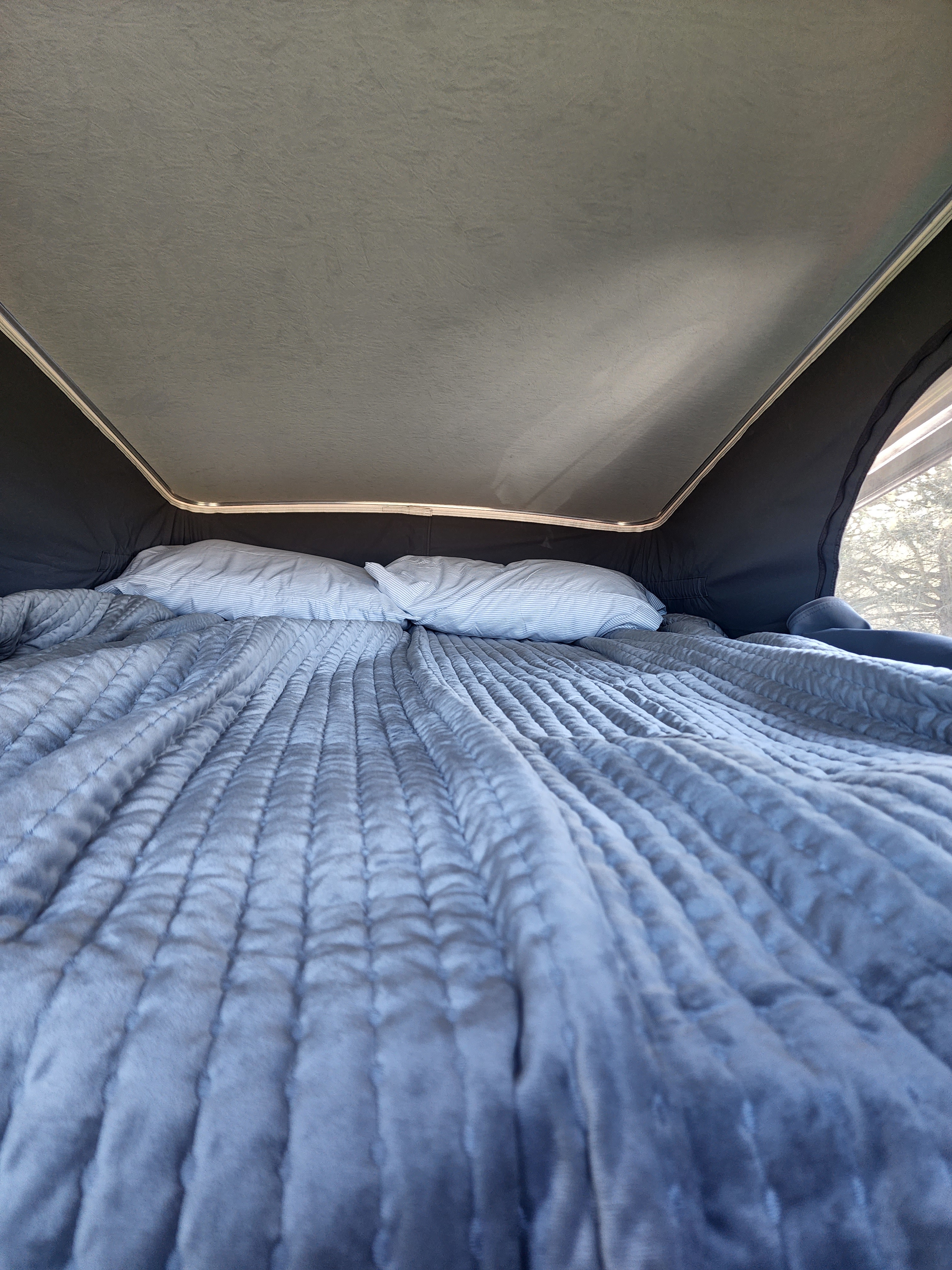 Cozy pop-top RV rental bed. A comfortable quilted grey blanket and two white pillows are ready for rest. Trees outside a mesh window.. Winnebago Solis 2022