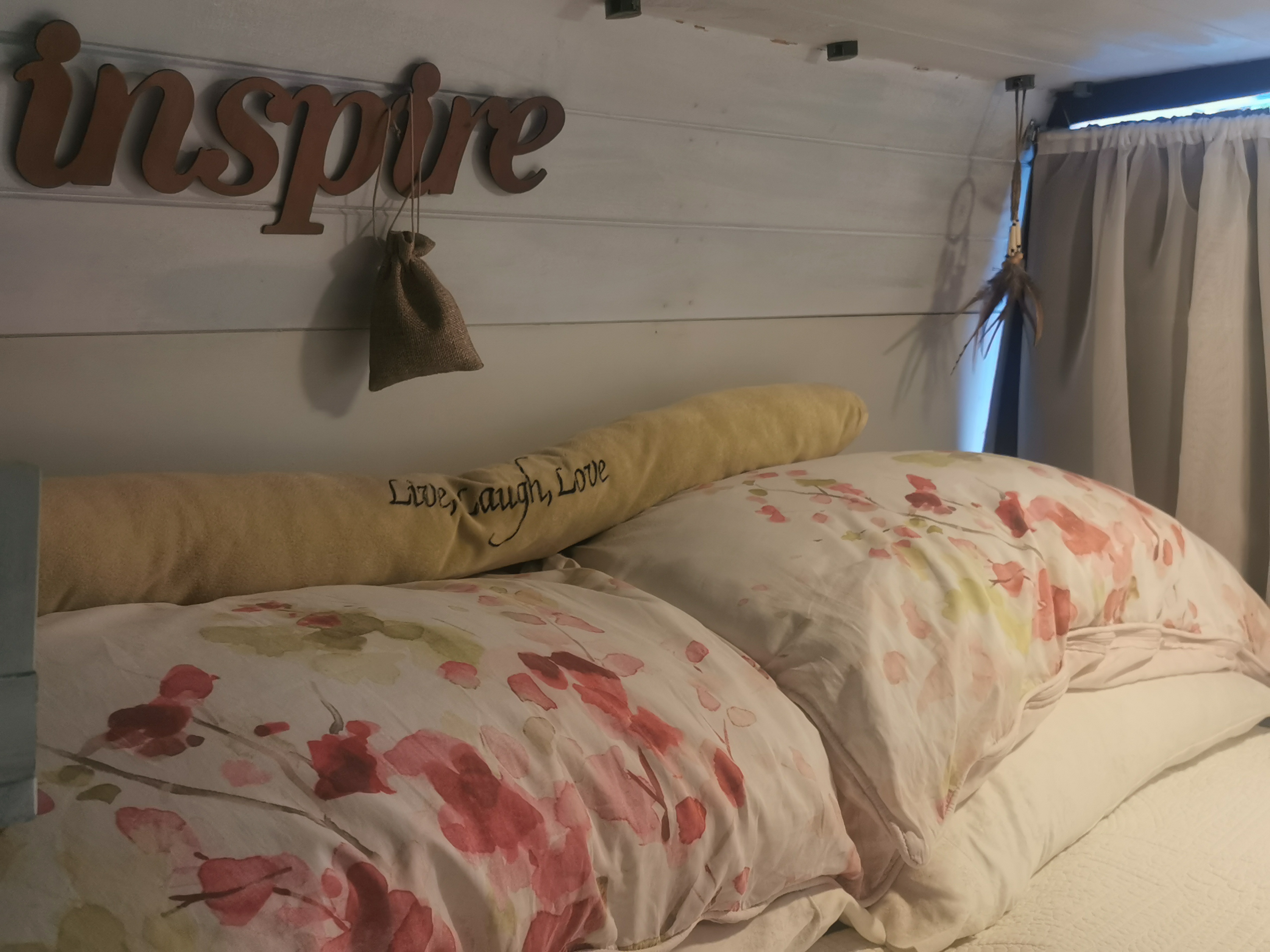 Cozy RV rental bedroom featuring an "inspire" sign, soft floral bedding, and a "Live, Laugh, Love" bolster pillow.. Dodge Sprinter 2006